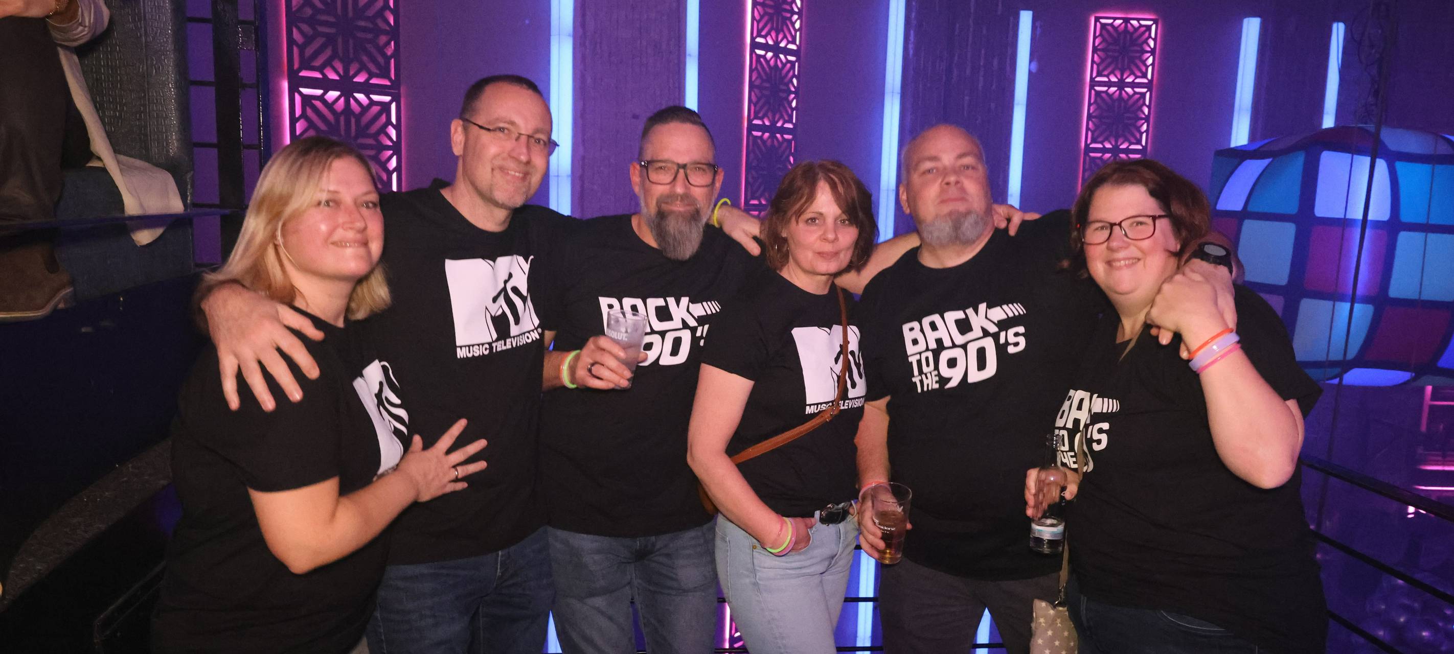 Drei Frauen und drei Männer in der Nachtresidenz beim Feiern. Sie liegen sich in den Armen. Alle tragen "back to the 90s" und "MTV"-T-Shirts.