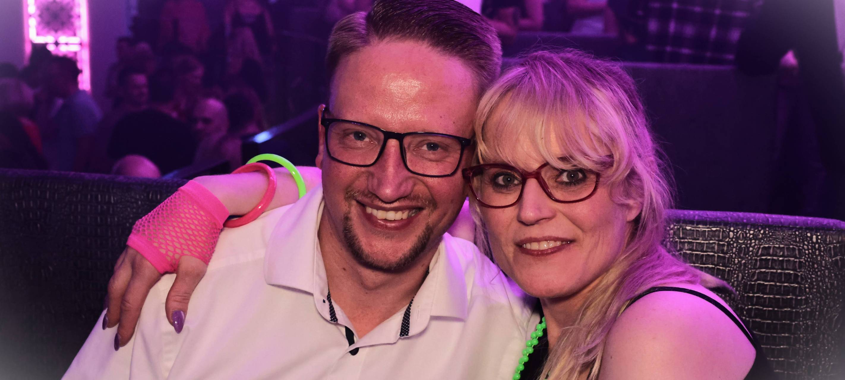 Ein Mann (links) und eine Frau (rechts) lassen sich gemeinsam in der Nachtresidenz fotografieren. Die Dame trägt Leuchtarmbänder. Im Hintergrund feiern Menschen.