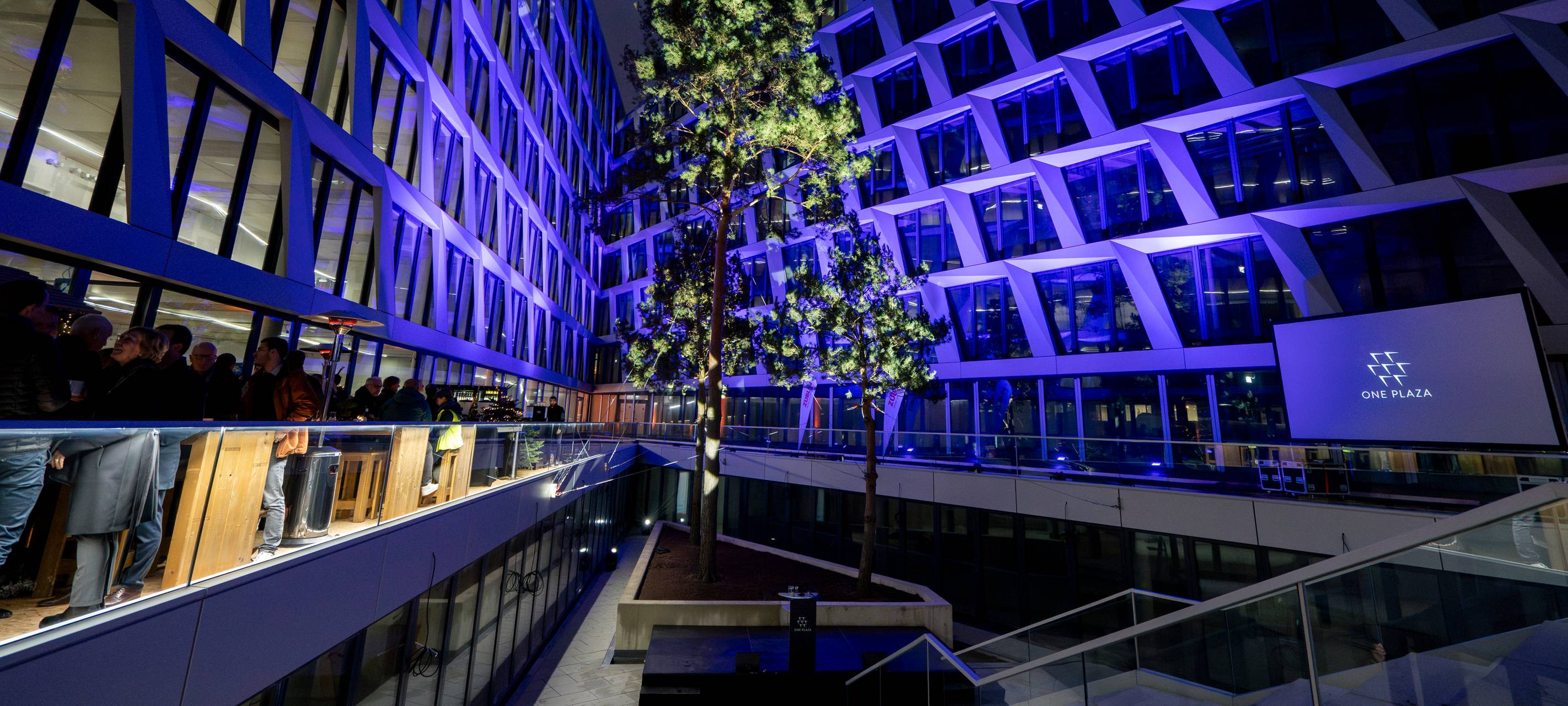 Das neue Bürogebäude One Plaza mit weißer Fassade und vielen Fenstern und der Innenhof mit drei hohen Bäumen.