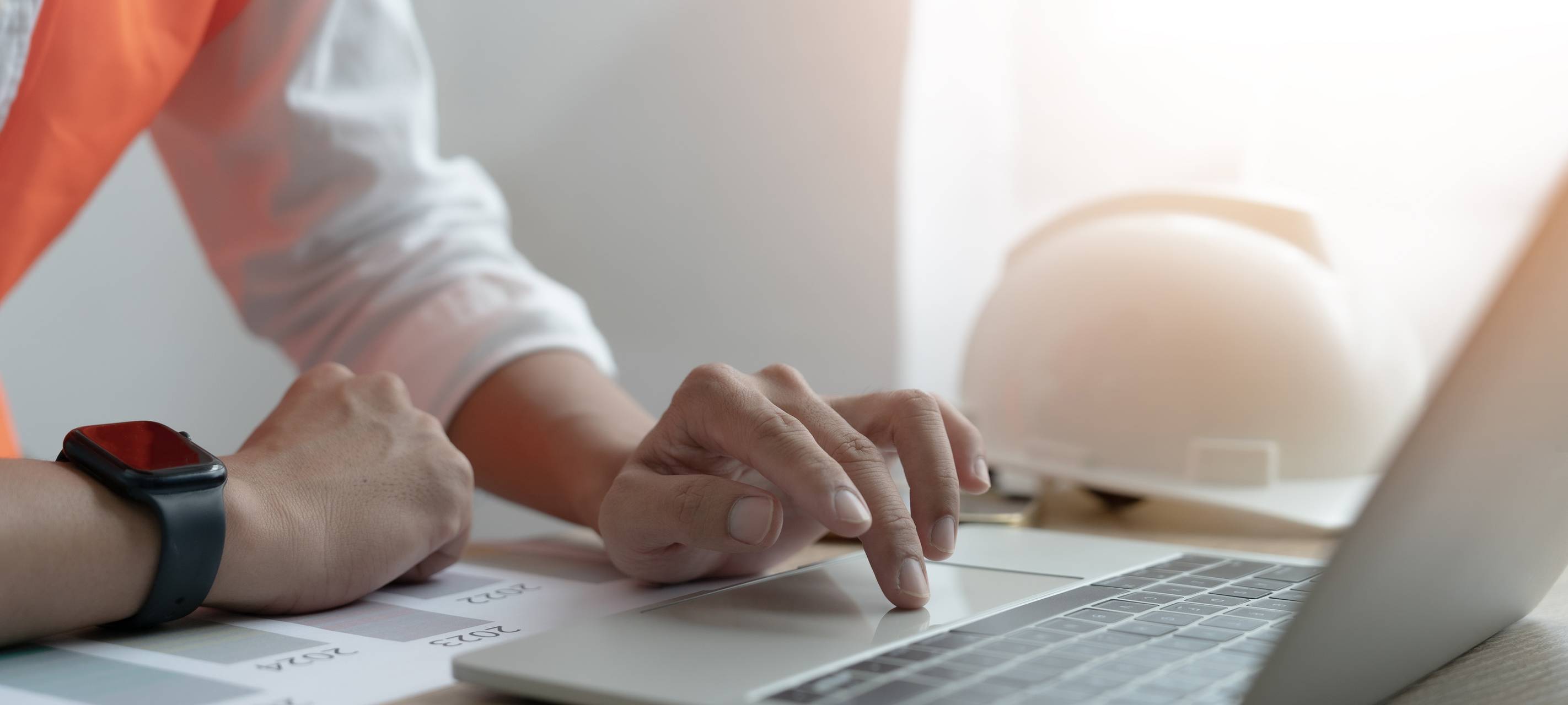 Eine Person tippt auf einem Laptop-Computer, während ein Bauhelm auf dem Schreibtisch liegt.