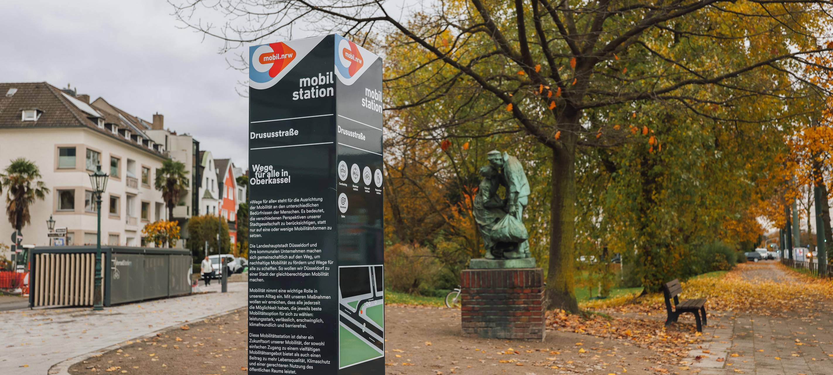 Eine Infostele weist auf die neue Mobilitätsstation an der Drususstraße in Oberkassel hin. Im Hintergrund sind Fahrradboxen zu erkennen.