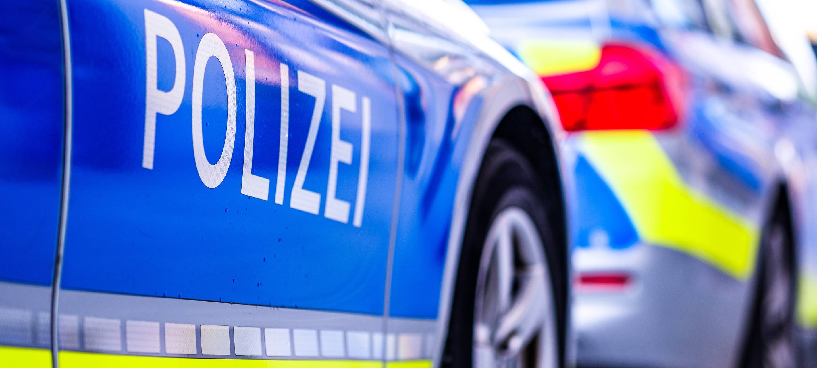 Ein Polizeiwagen auf einer Straße in Deutschland. Ein Polizeiwagen wird von der Seite abgebildet. Er ist blau-weiß mit gelben Streifen und in weißen Großbuchstaben steht "Polizei".