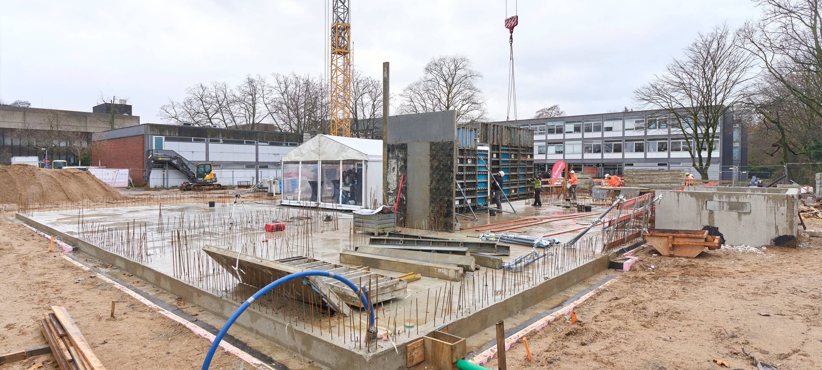 Baustelle für die Erweiterung der Rolandschule. Rund um den Grundstein befinden sich eine große Betonplatte, Kräne, Rohre und Kabel.