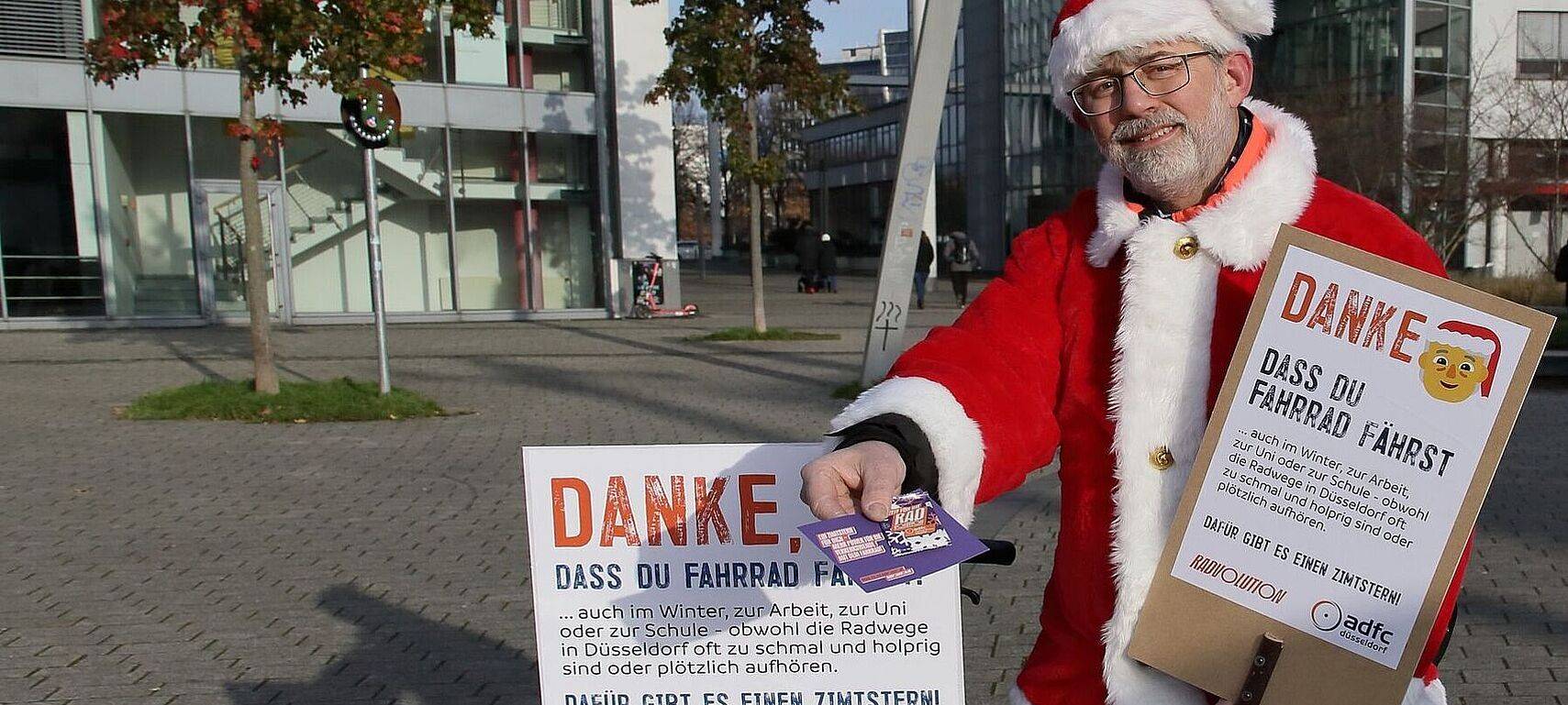 Ein verkleideter Nikolaus hält der Kamera einen Flyer und eine Zimtschnecke entgegen. In seiner Hand hält er ein Schild mit dem Slogan "Danke, dass du Fahrrad fährst!".