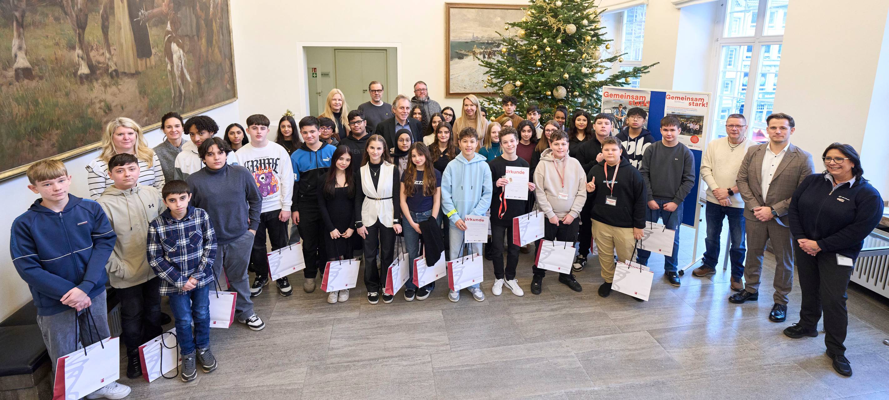 Neue Fahrzeugbegleiter für mehr Sicherheit haben sich im Düsseldorfer Rathaus versammelt. Sie stehen in einer Reihe mit ihren Urkunden und Taschen im Jan-Wellem-Saal vor einem Weihnachtsbaum.