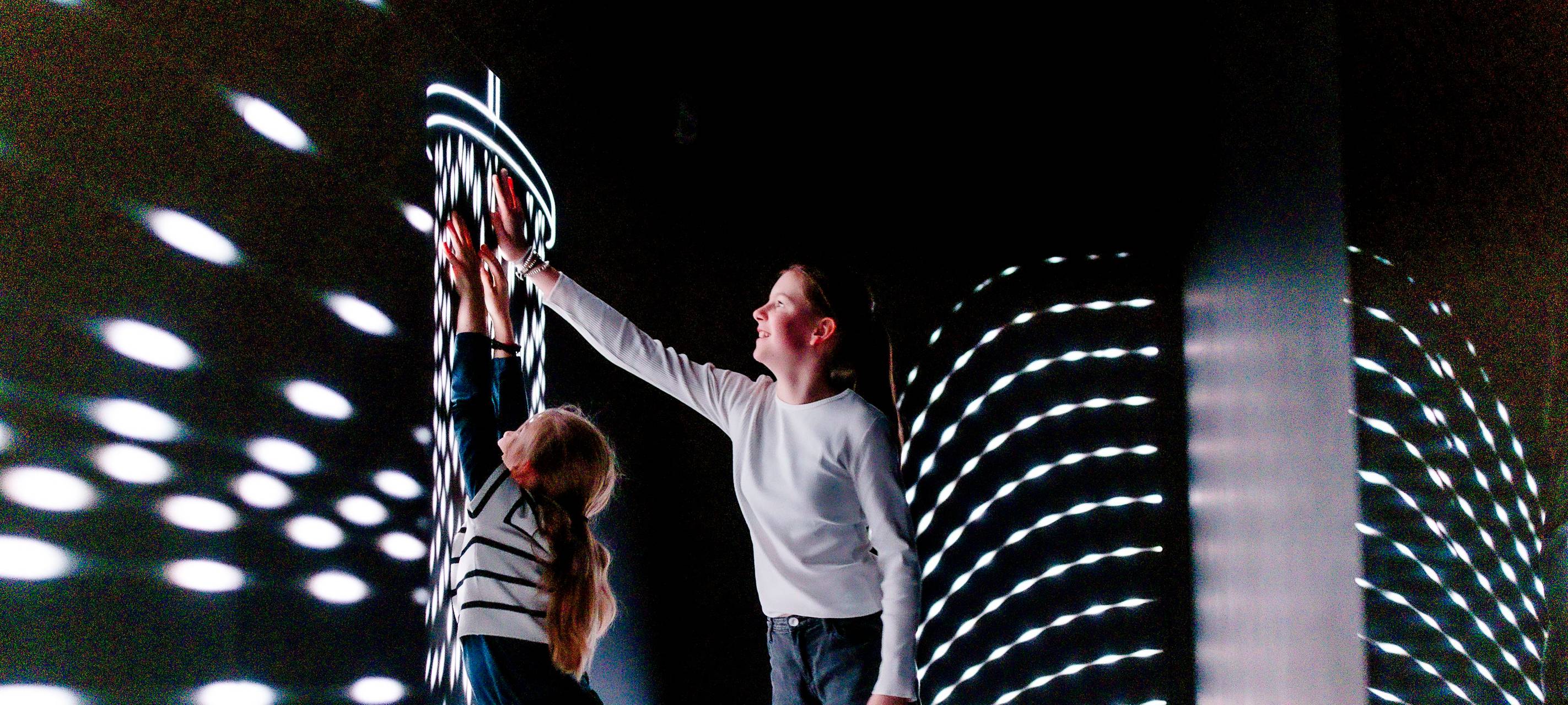 Zwei Kinder stehen im Kinderraum mit einer besonderen Lichtinstallation im Düsseldorfer Kunstpalast. Beide Kinder strecken sich und fassen eine Wand an.