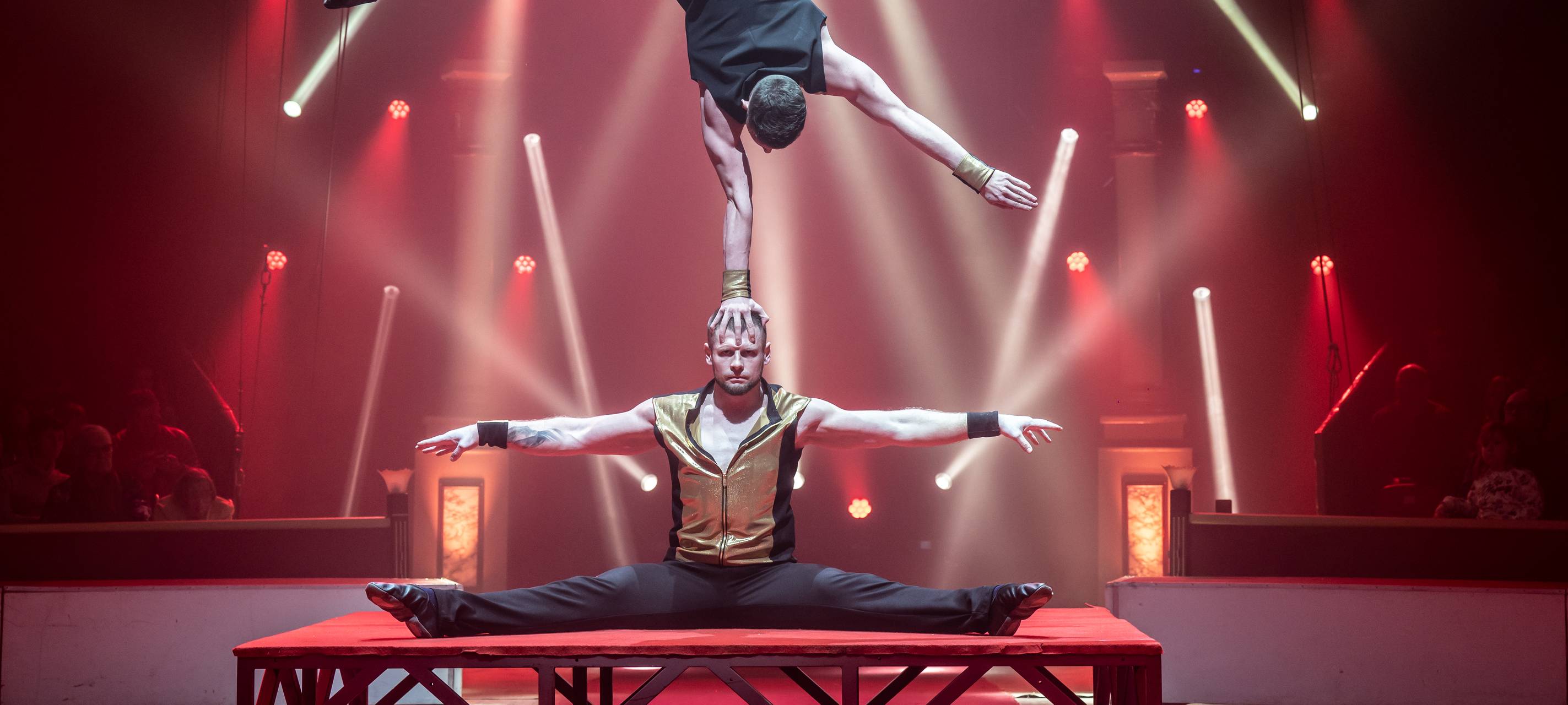 Ein Artist sitzt im Spagat auf dem Boden und streck die Arme aus, ein weiterer balanciert mit einer Hand auf seinem Kopf