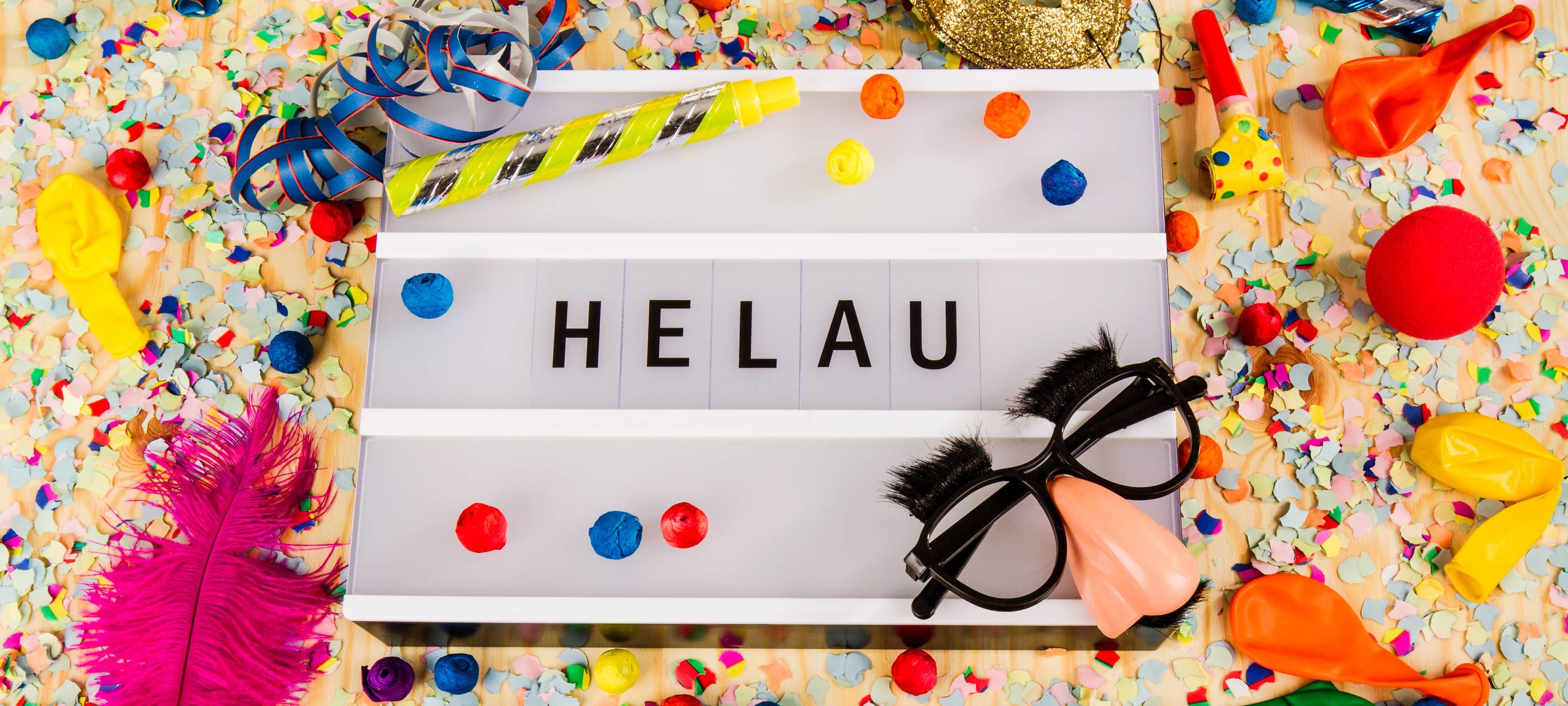 Ein festliches Szenario mit Konfetti, Luftballons, Partytröten, einer goldenen Maske und Spaßbrillen umgibt eine Leuchtbox mit der Aufschrift "HELAU."