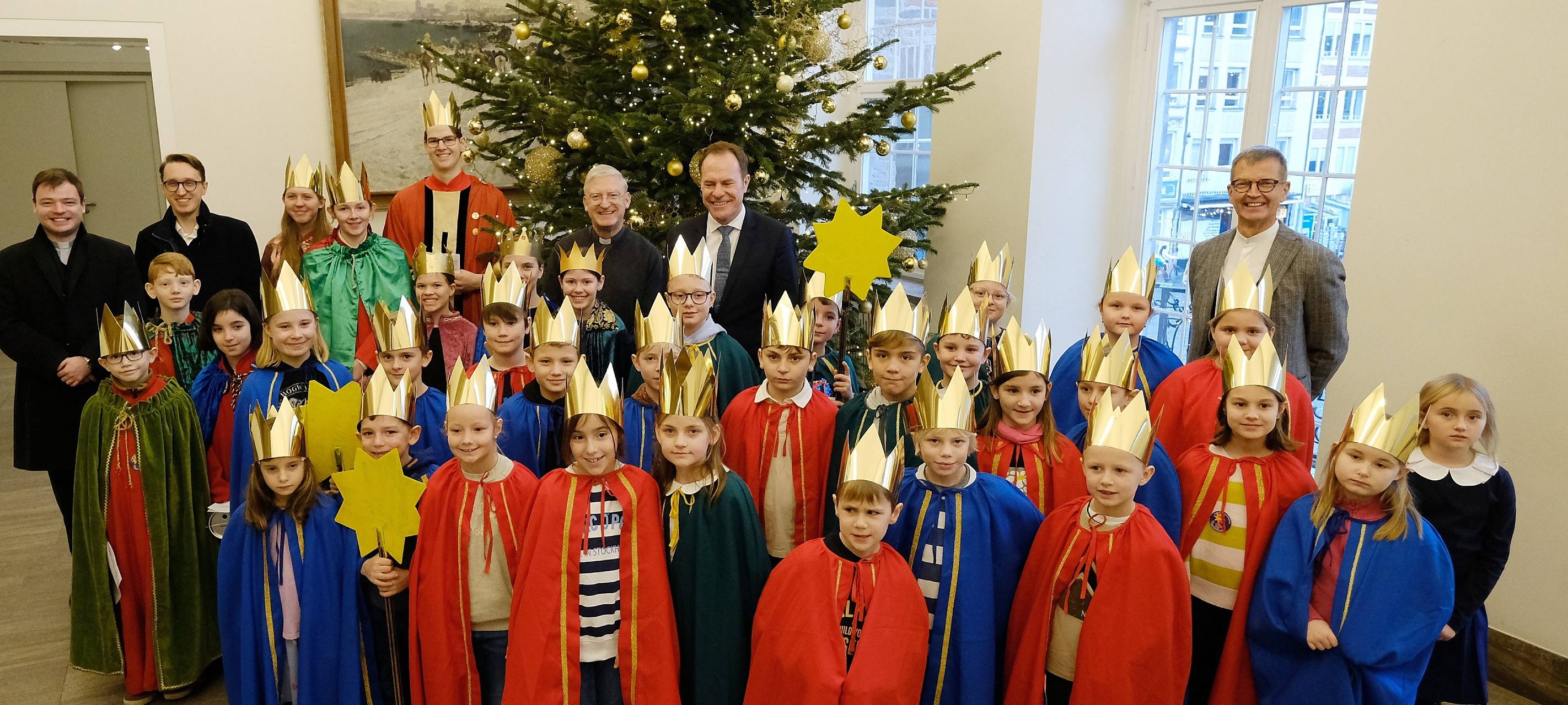 Rund 30 Kinder stehen als Sternsinger vor dem Tannenbaum im Rathaus. Sie tragen gebastelte goldene Kronen. Einige halten außerdem einen Stern in der Hand.