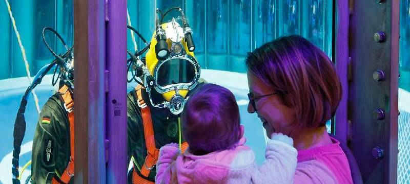 Eine Frau schaut mit ihrem Kind auf dem Arm auf einen Taucher, der hinter einer Scheibe im Tauchbecken ist.