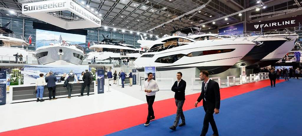 Messe boot: Drei Männer laufen im Vordergrund auf einem blau-roten Teppich durch die Messehalle. Sie unterhalten sich. Im Hintergrund kann man große Yachten und weiteres Publikum erkennen.