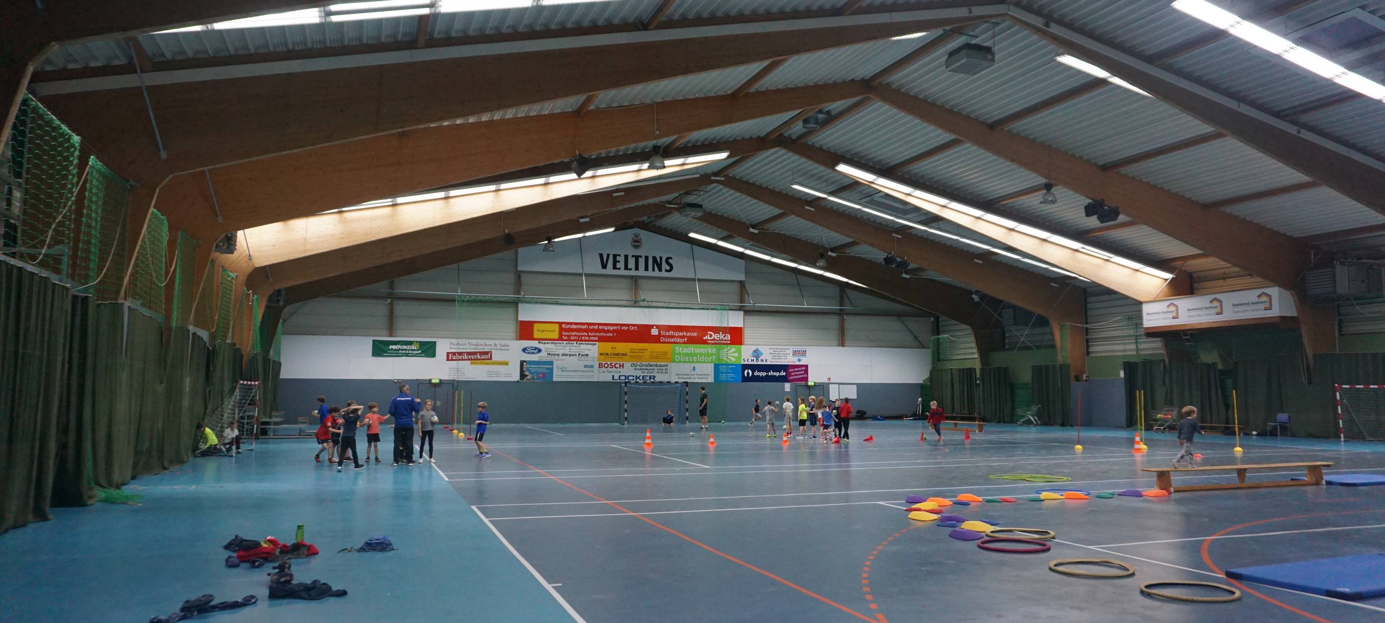 Zu sehen ist die Mehrzwecksporthalle in Düsseldorf Angermund. Im Hintergrund sieht man spielende Kinder, die an einem Training teilnehmen.