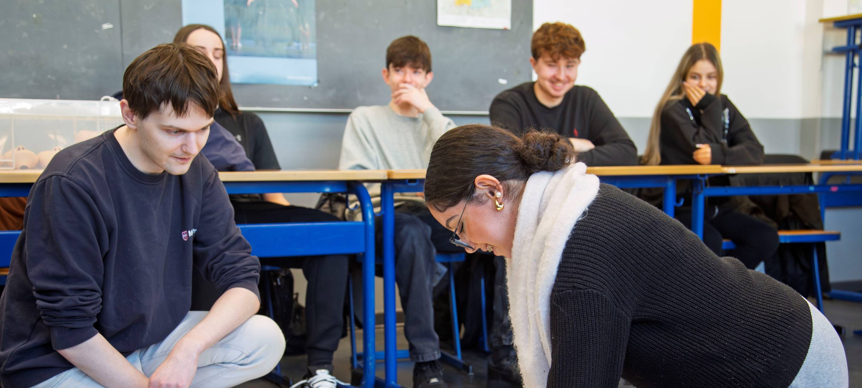 Eine Schülerin und ein Schüler üben die Wiederbelebung an einer Puppe. Im Hintergrund sitzen weitere Schüler.