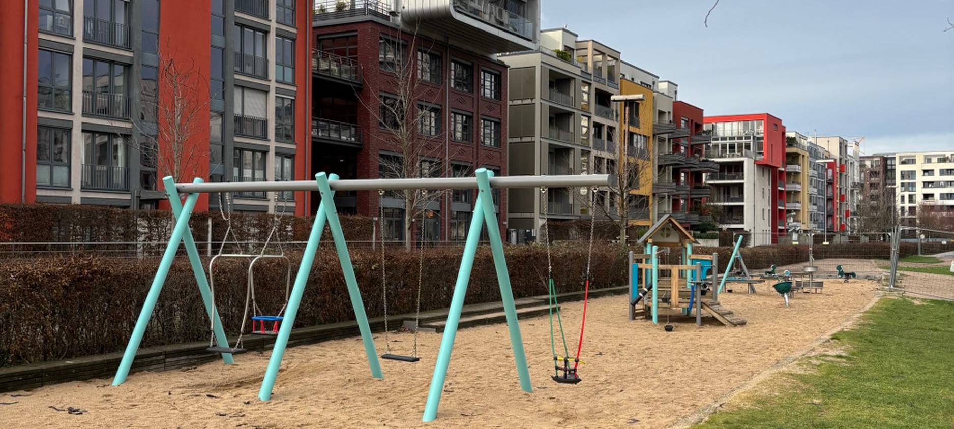 Eine Spielplatz, bestehend aus einer Sandfläche mit Spielgeräten. Im Vordergrund sind Schaukeln. Hinter dem Spielplatz stehen Mehrfamilienhäuser.