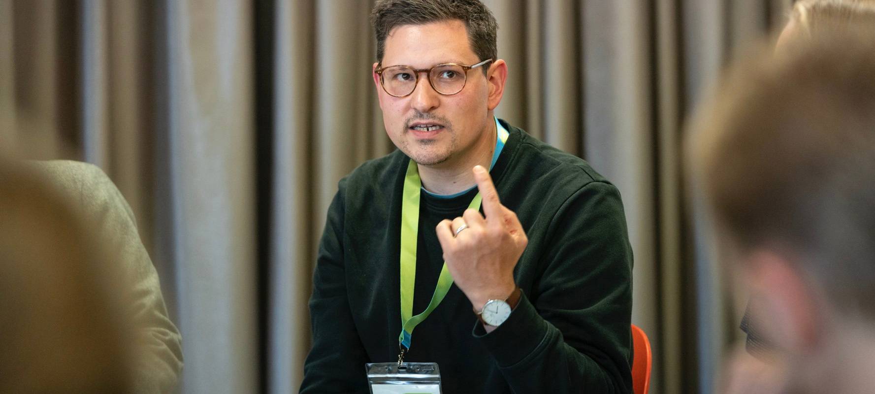 Ein Mann (Dennis Frieß) sitzt mit erhobenem Zeigefinger vor einem Vorhang und hält einen Vortrag. Im Vordergrund sind andere Menschen zu erahnen.