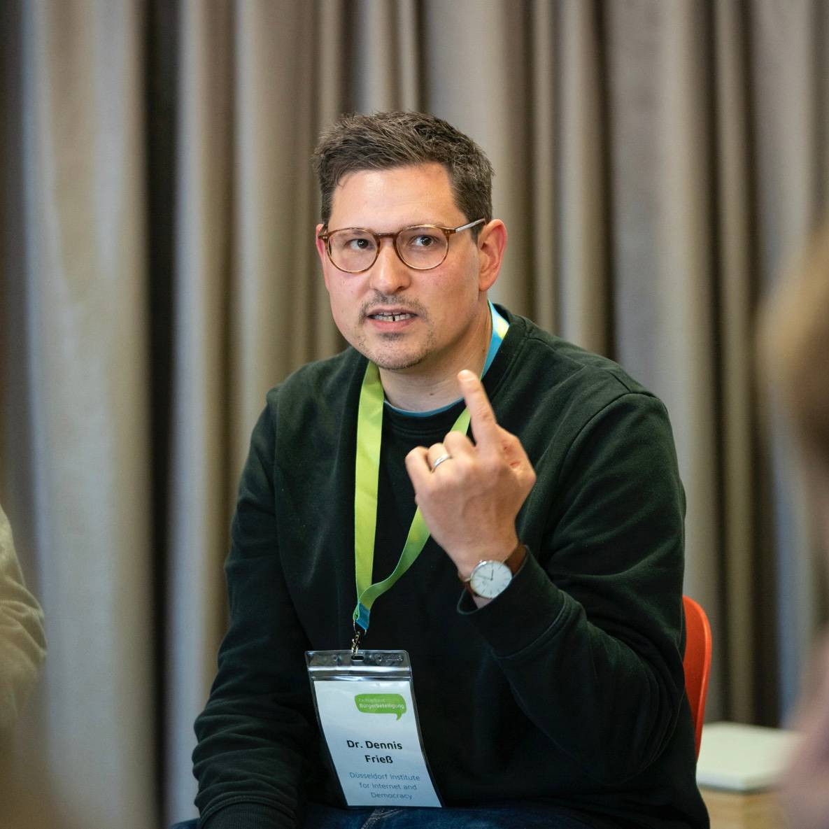 Ein Mann (Dennis Frieß) sitzt mit erhobenem Zeigefinger vor einem Vorhang und hält einen Vortrag. Im Vordergrund sind andere Menschen zu erahnen.