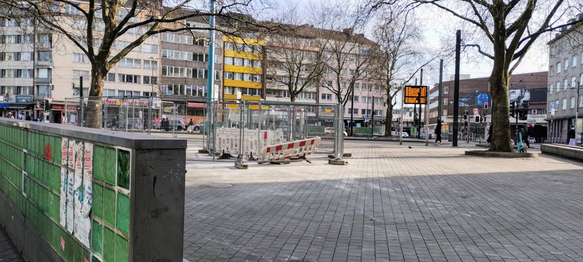 Worringer Platz in Düsseldorf nach dem Aufräumen. In der Mitte stehen Bauzäune. Am Rand des Platzes stehen grüne Glasbausteine. Rundherum gibt es einige Bäume.