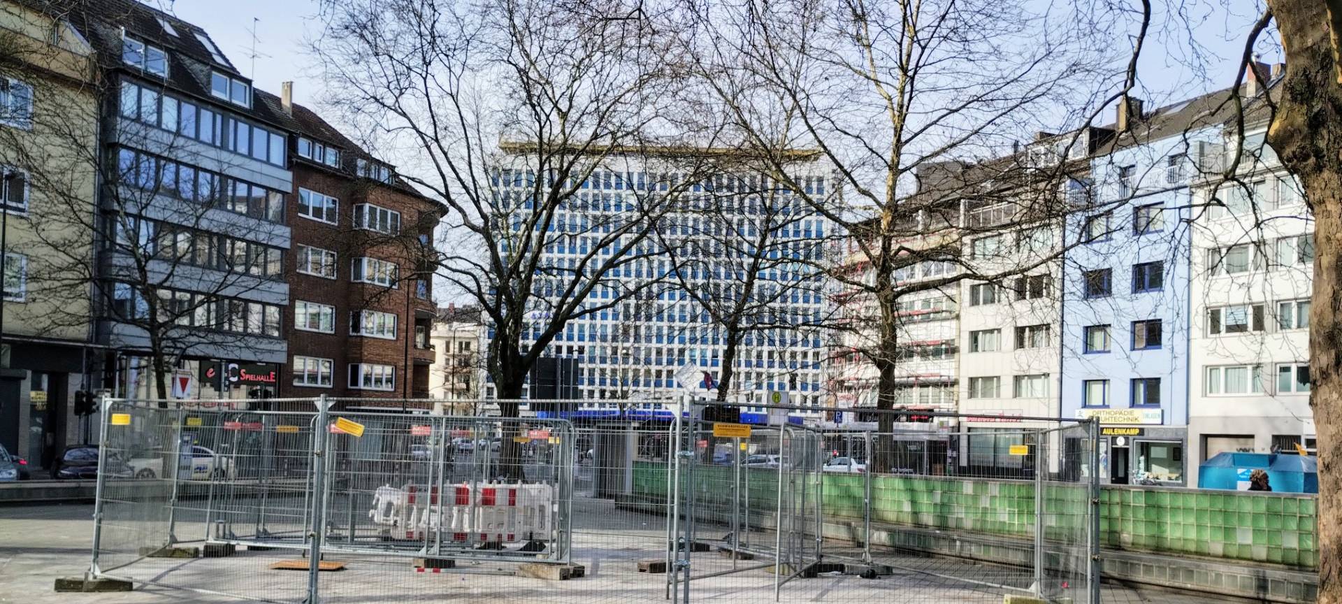 Worringer Platz in Düsseldorf nach dem Aufräumen. In der Mitte stehen Bauzäune. Am Rand des Platzes stehen grüne Glasbausteine. Rundherum gibt es einige Bäume.