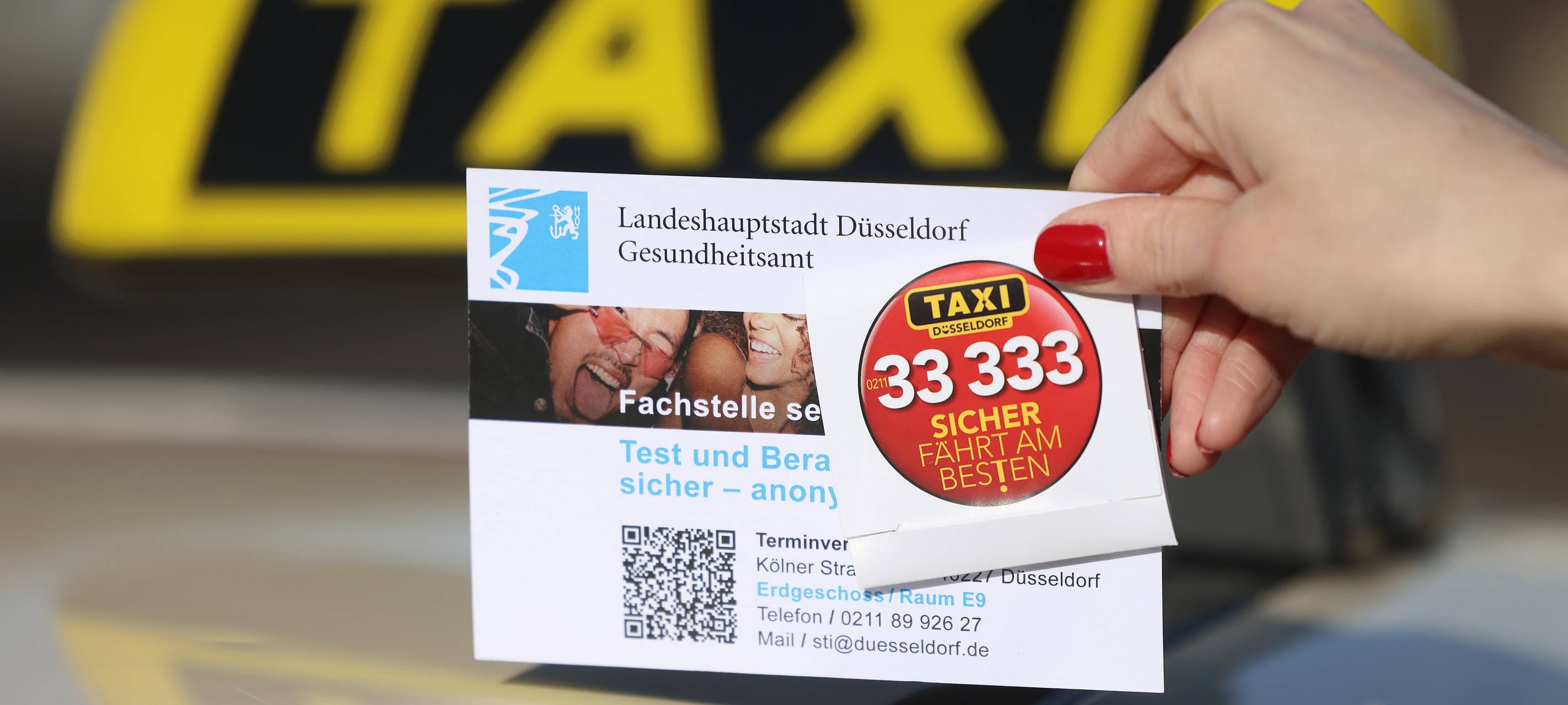 Foto zur Kondomaktion der Taxi-Innung Düsseldorf. Eine Frau hält ein Werbeschild mit Kondom vor einem Taxi-Schild.