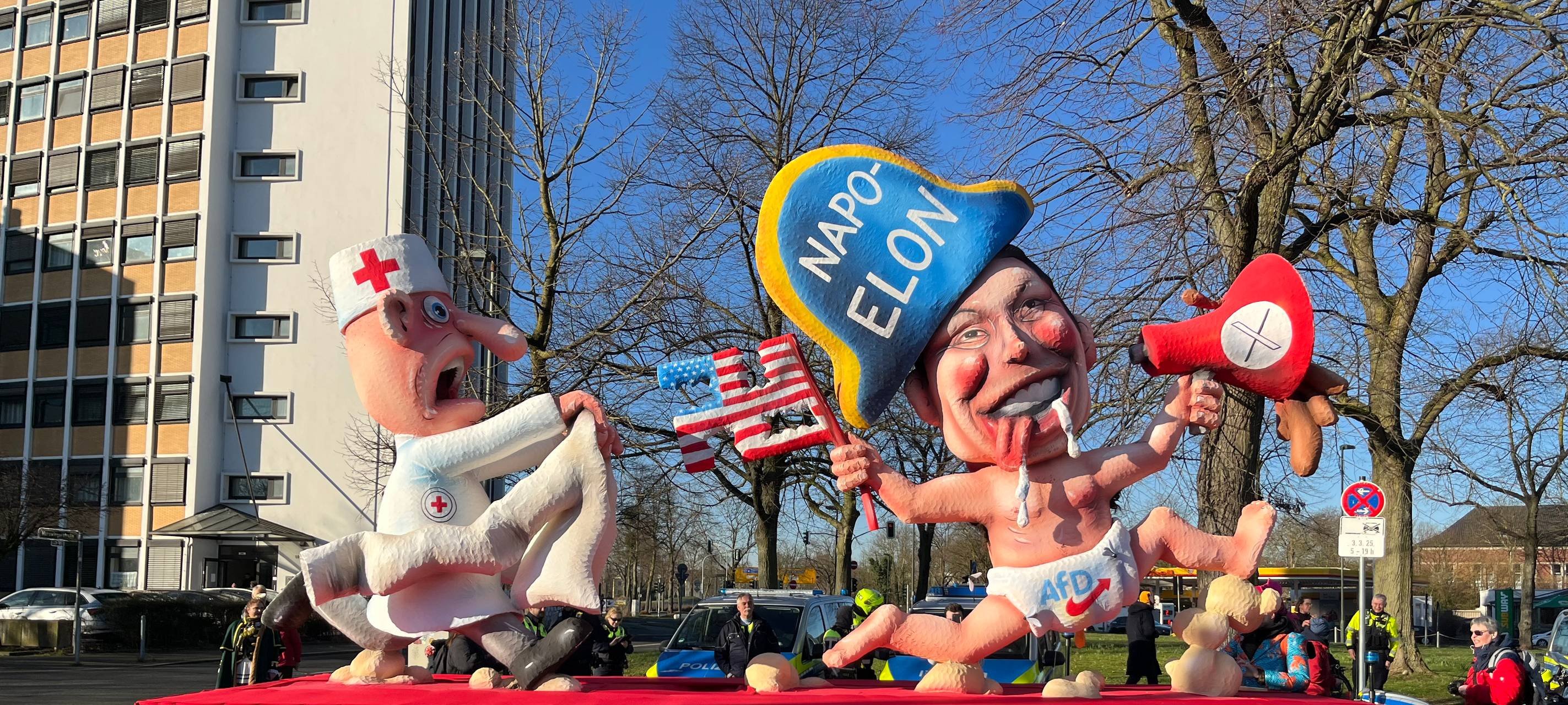 Mottowagen beim Karneval: Elon Musk als Napolon in AfD-Windel wird von einem Arzt mit Zwangsjacke verfolgt.