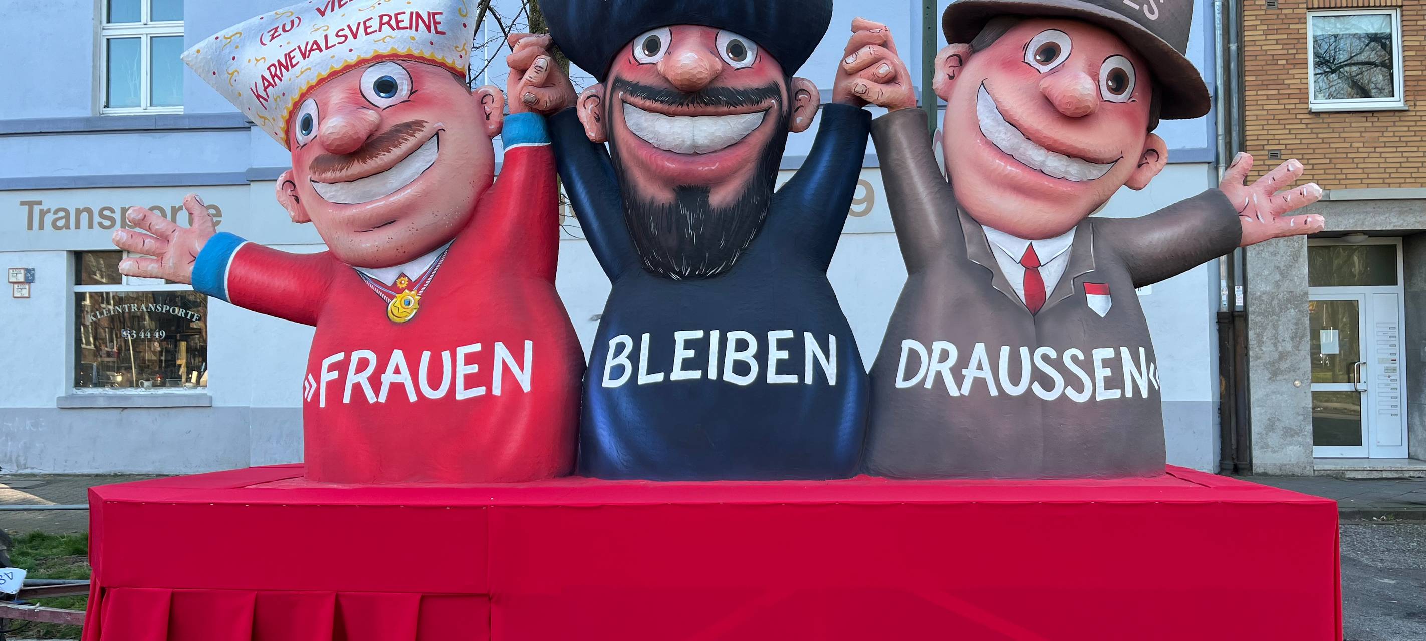 Mottowagen beim Karneval: Karnevalsvereine, Mullahs und die Düsseldorfer Jonges halten sich an den Händen und reißen die Arme hoch. Vorne steht "Frauen müssen draußen bleiben".