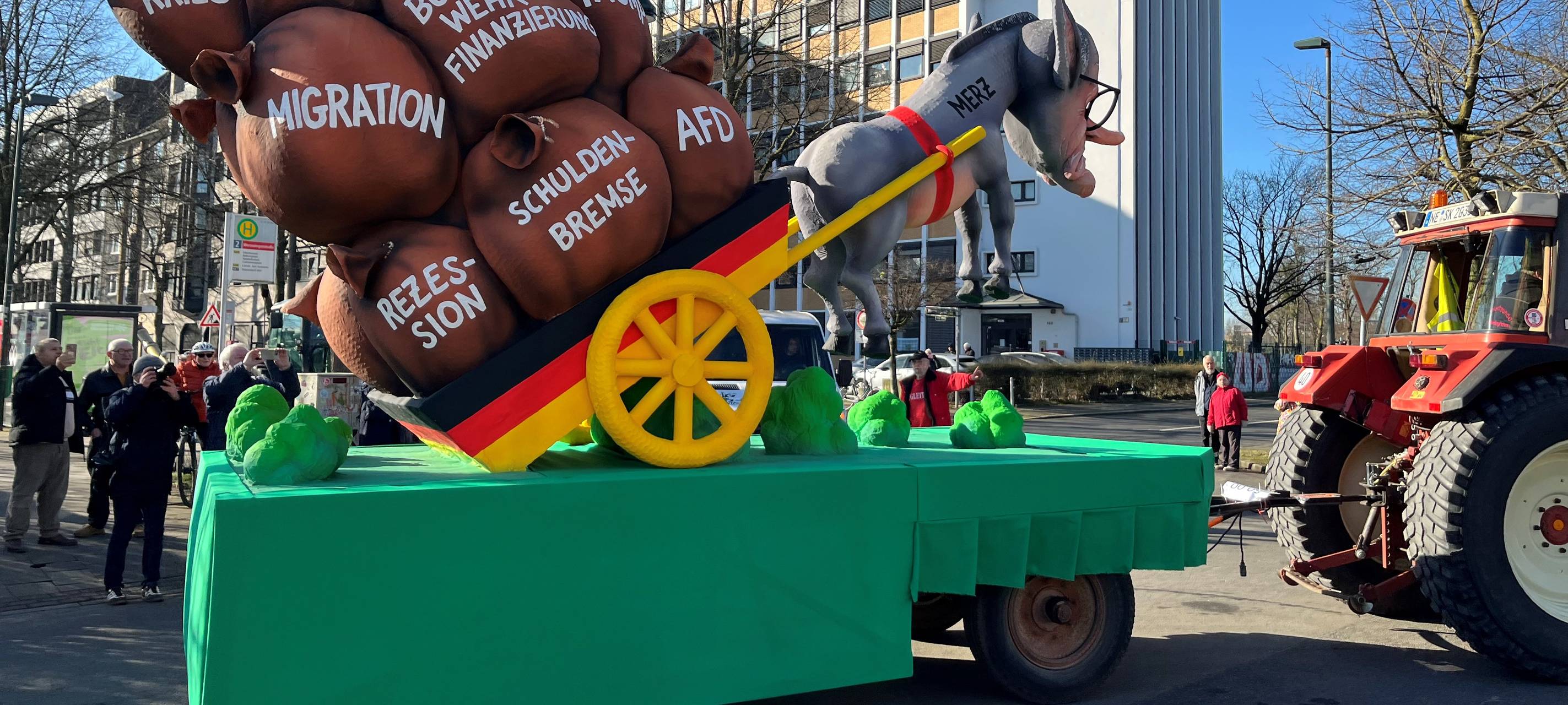 Mottowagen beim Karneval: Merz als Esel vor dem Deutschland-Karren hängt in der Luft, Auf dem Karren liegen Säcke mit Aufschriften wie "AfD", "Migration" oder "Schuldenbremse"
