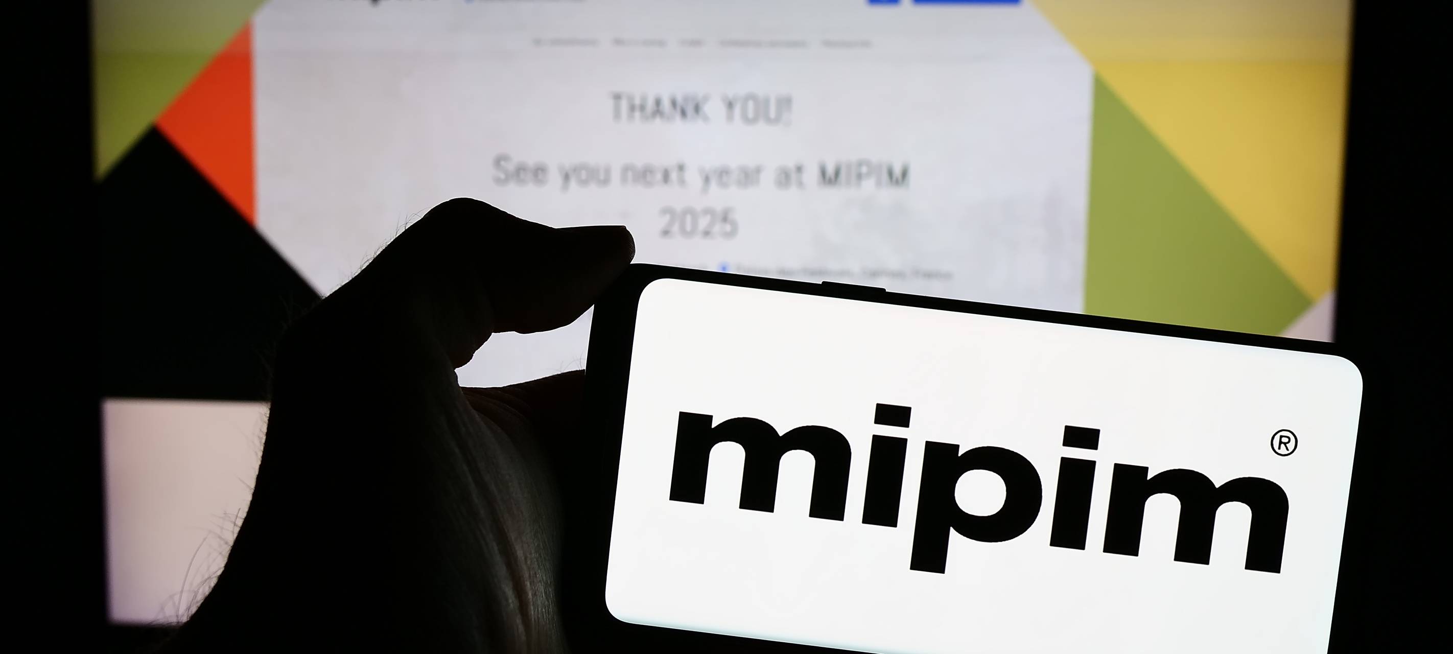 Person, die ein Mobiltelefon mit dem Logo der französischen Immobilienkonferenz MIPIM vor einer Geschäftswebseite hält. Fokus auf dem Display des Telefons.
