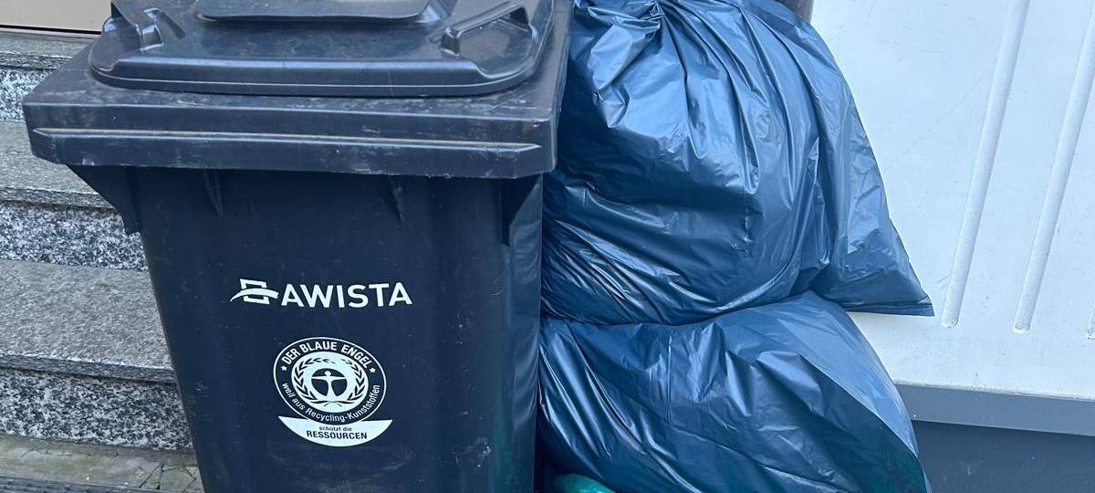 Eine Restmülltonne steht vor einer Hauswand, daneben mehrere verschlossene Säcke mit Restmüll.