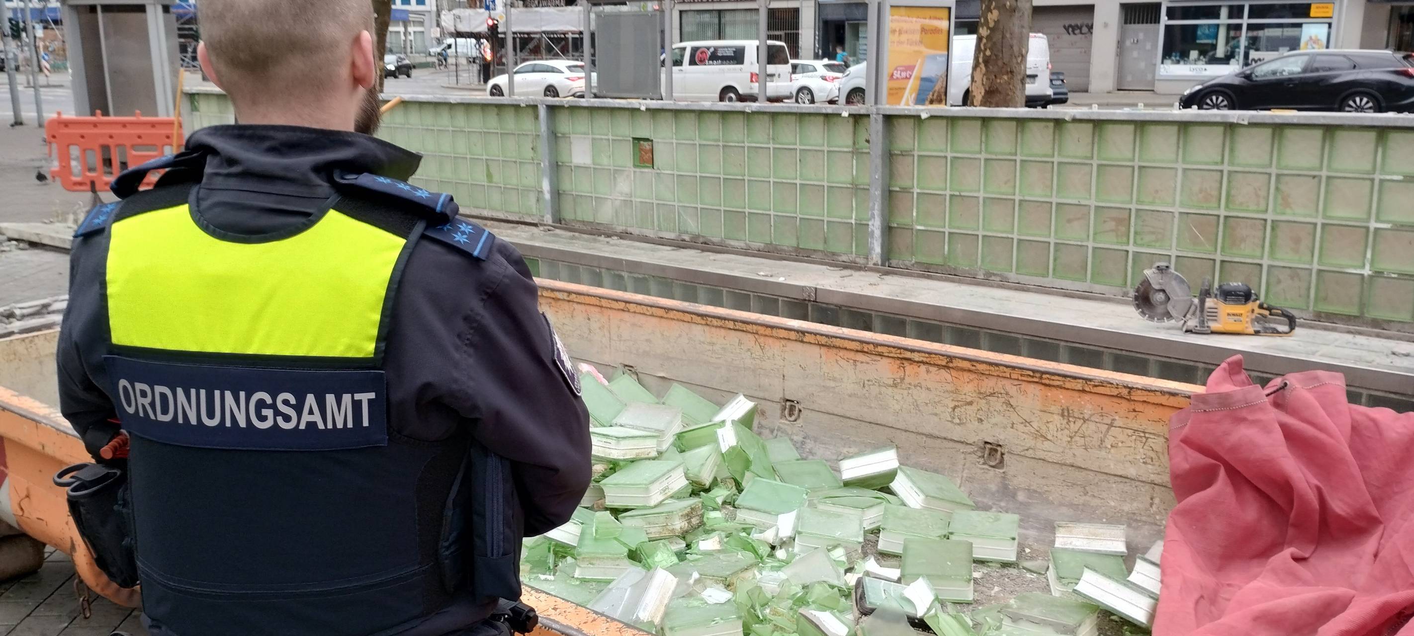 Ein Mitarbeiter des Ordnungsamtes steht vor einem Container mit herausgeschlagenen Glasbausteinen. Im Hintergrund die grünen Bänke am Worringer Platz, die jetzt nach und nach entfernt werden.