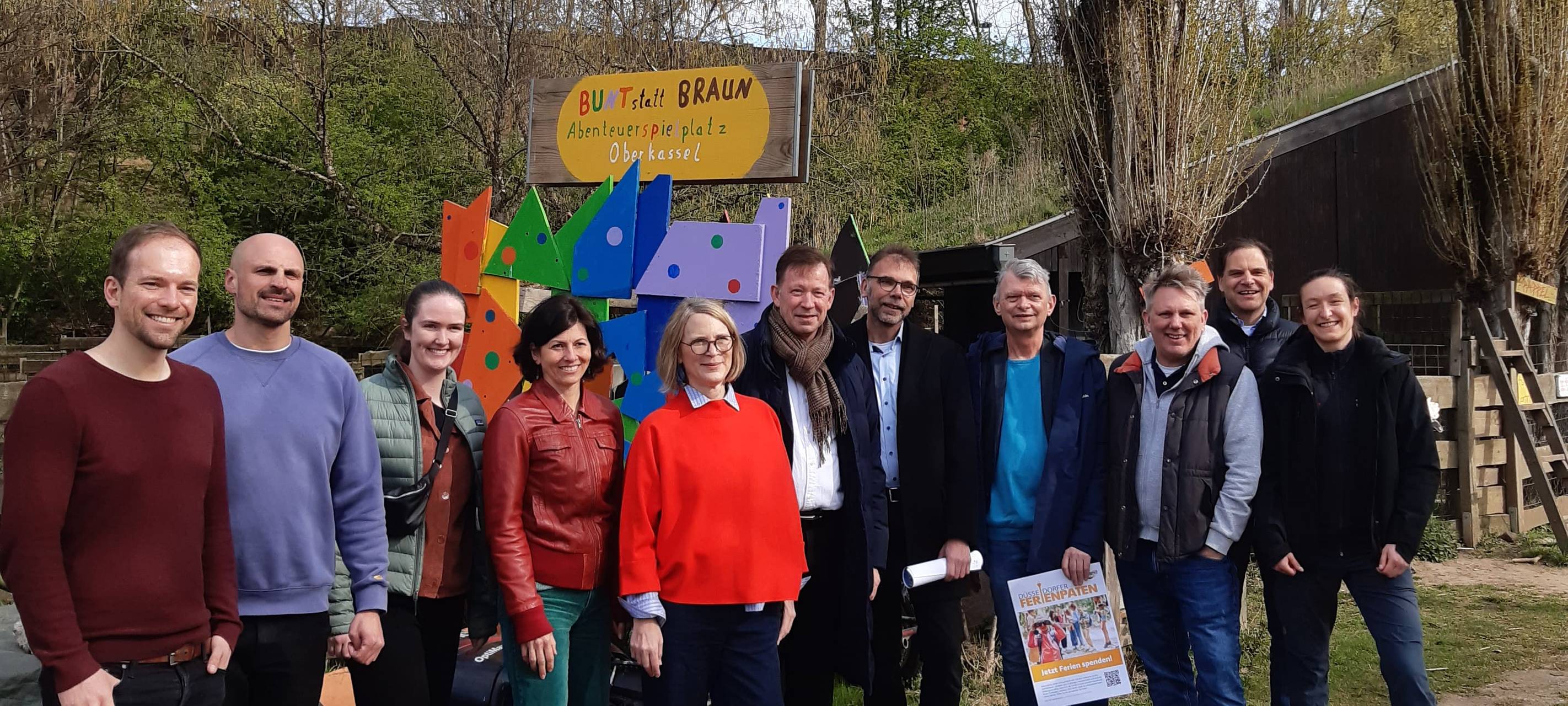 Ein Menschengruppe hat sich vor einem bunten Schild aufgestellt, alle Lächeln in die Kamera. Im Hintergrund sind Bäume in Sträucher.