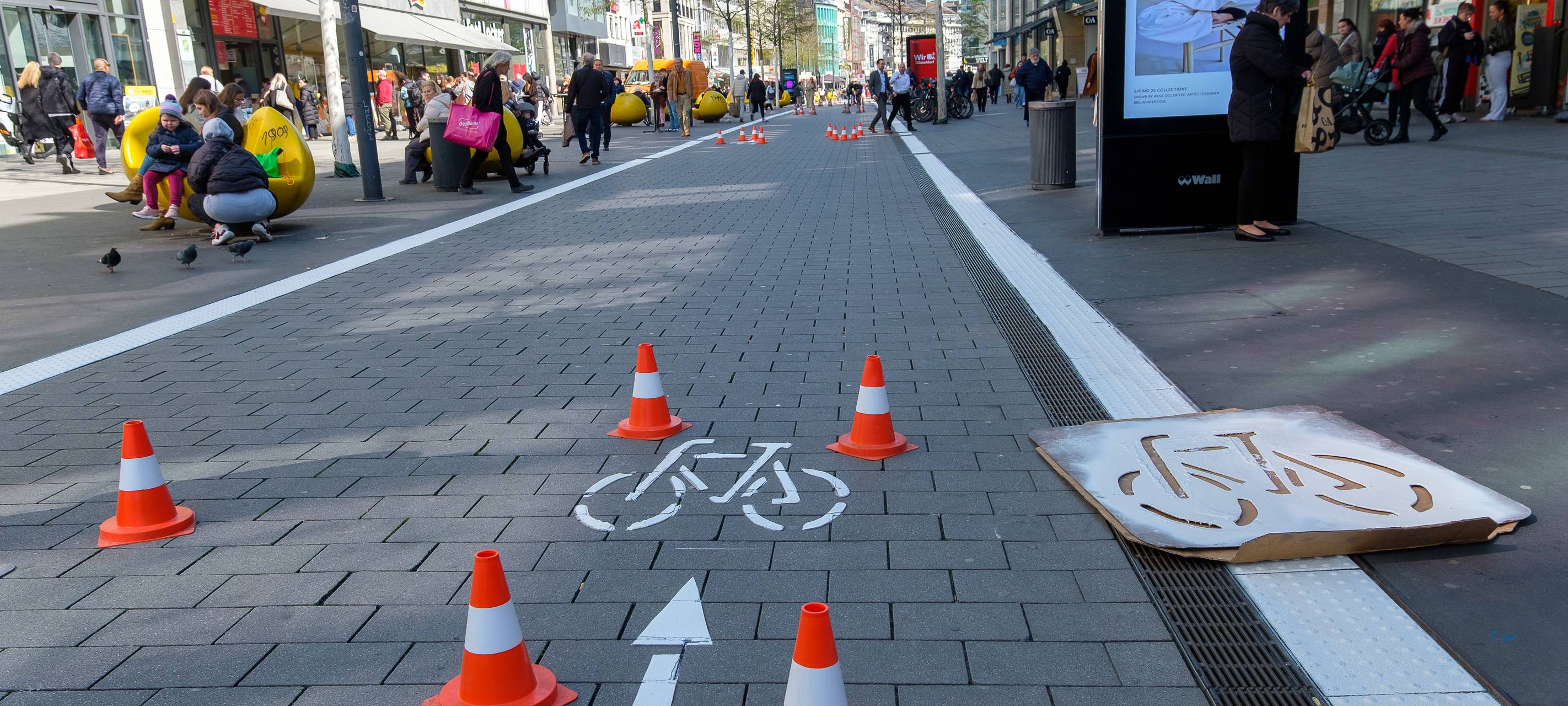 Der Radweg auf der Schadowstraße ist mit weißer Farbe teilweise neu markiert. Zu sehen ist unter anderem ein Fahrrad-Piktogramm. Links und rechts vor den Geschäftshäusern sind Menschen zu Fuß unterwegs.