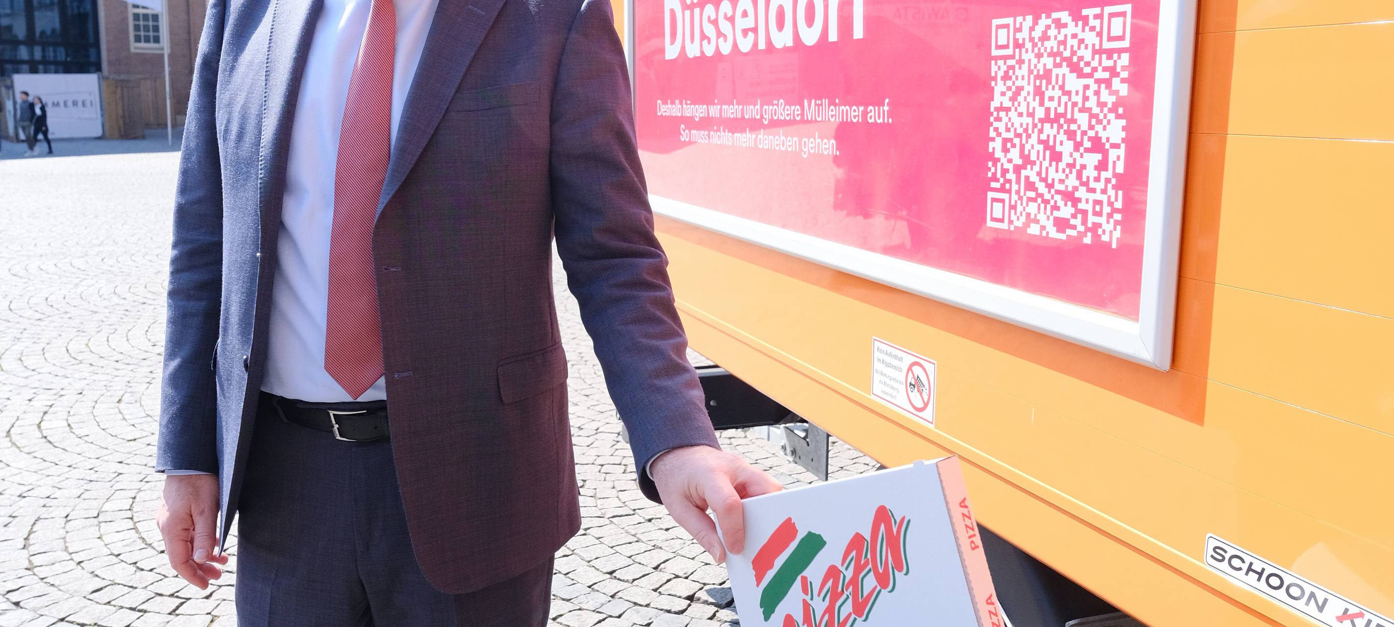 OB Keller wirft vor dem Rathaus einen Pizzakarton in einen Mülleimer mit extra breitem Schlitz. Daneben ist ein AWISTA-Fahrzeug zu sehen.