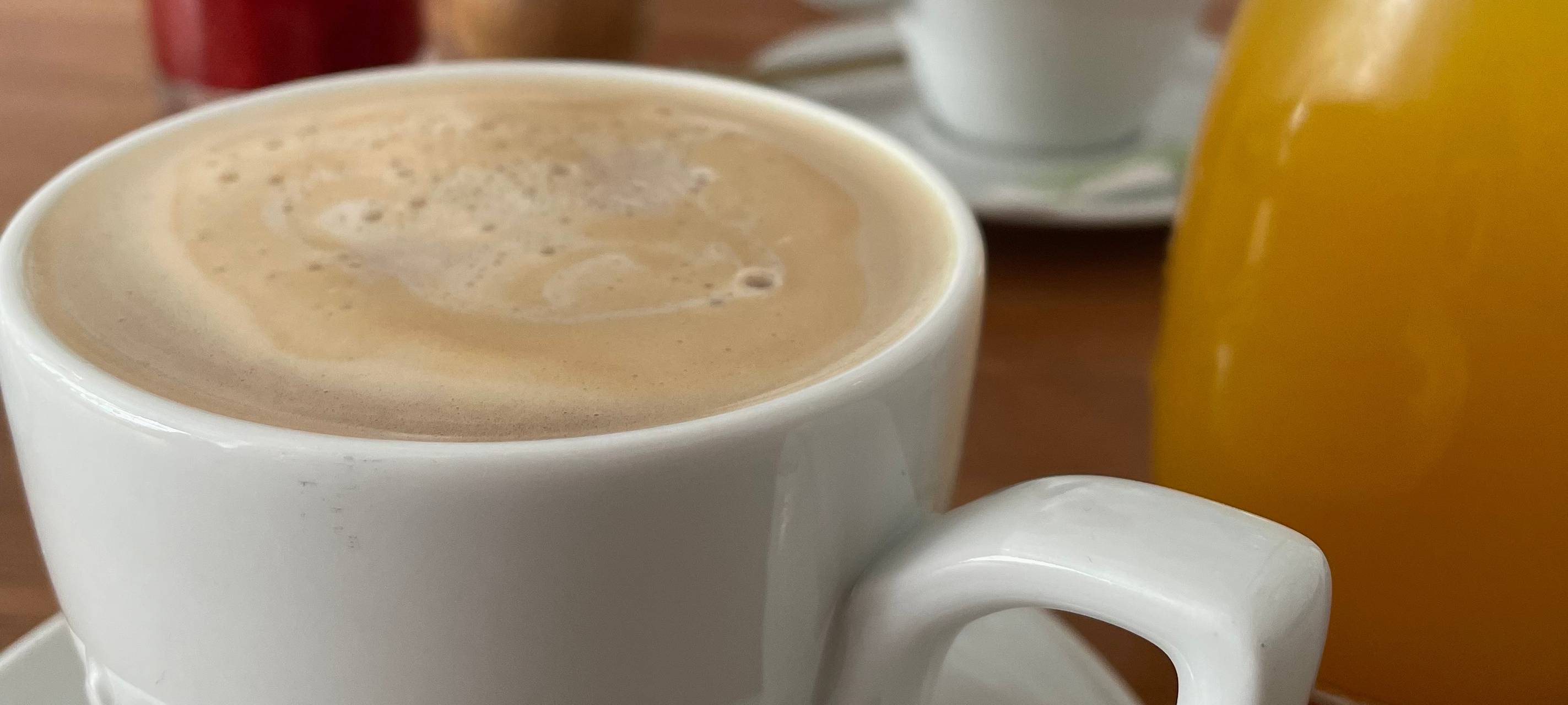 Eine Tasse Kaffee, daneben ein Glas mit einem Saft. Auf dem Tisch im Hintergrund steht eine Kerze.