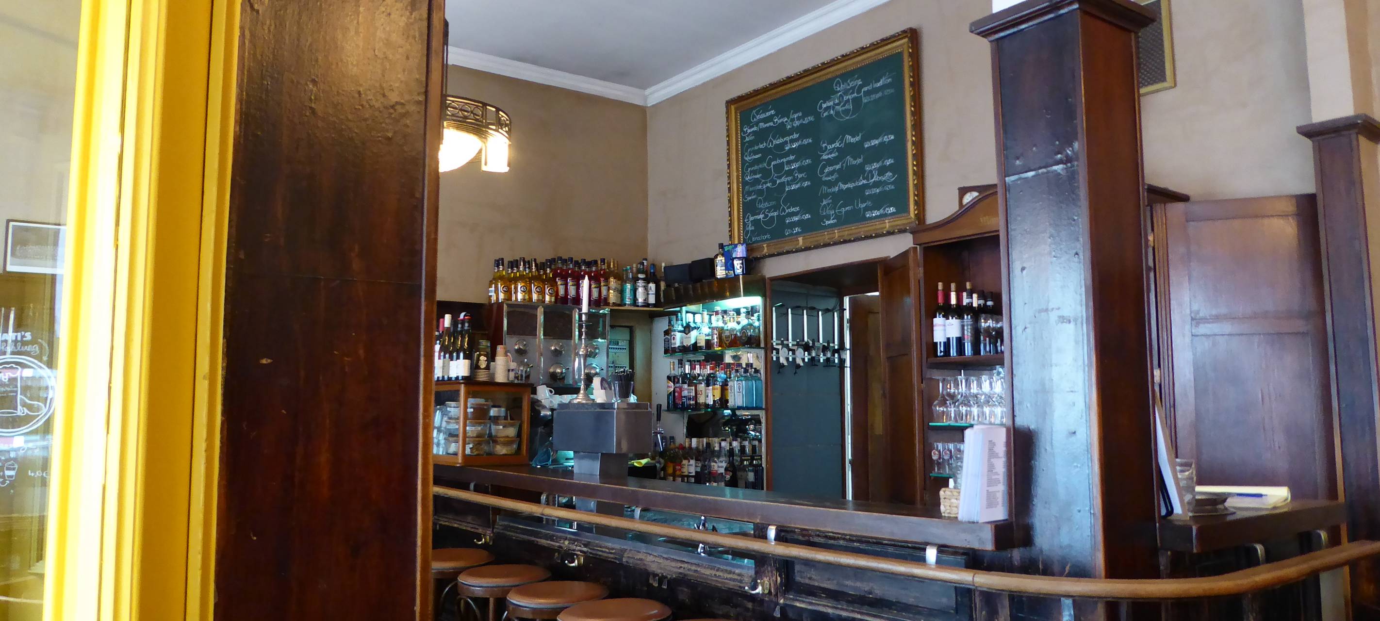 Links ein massiver Holzbalken, dahinter eine dunkelbraune Theke, vor der Barhocker stehen.