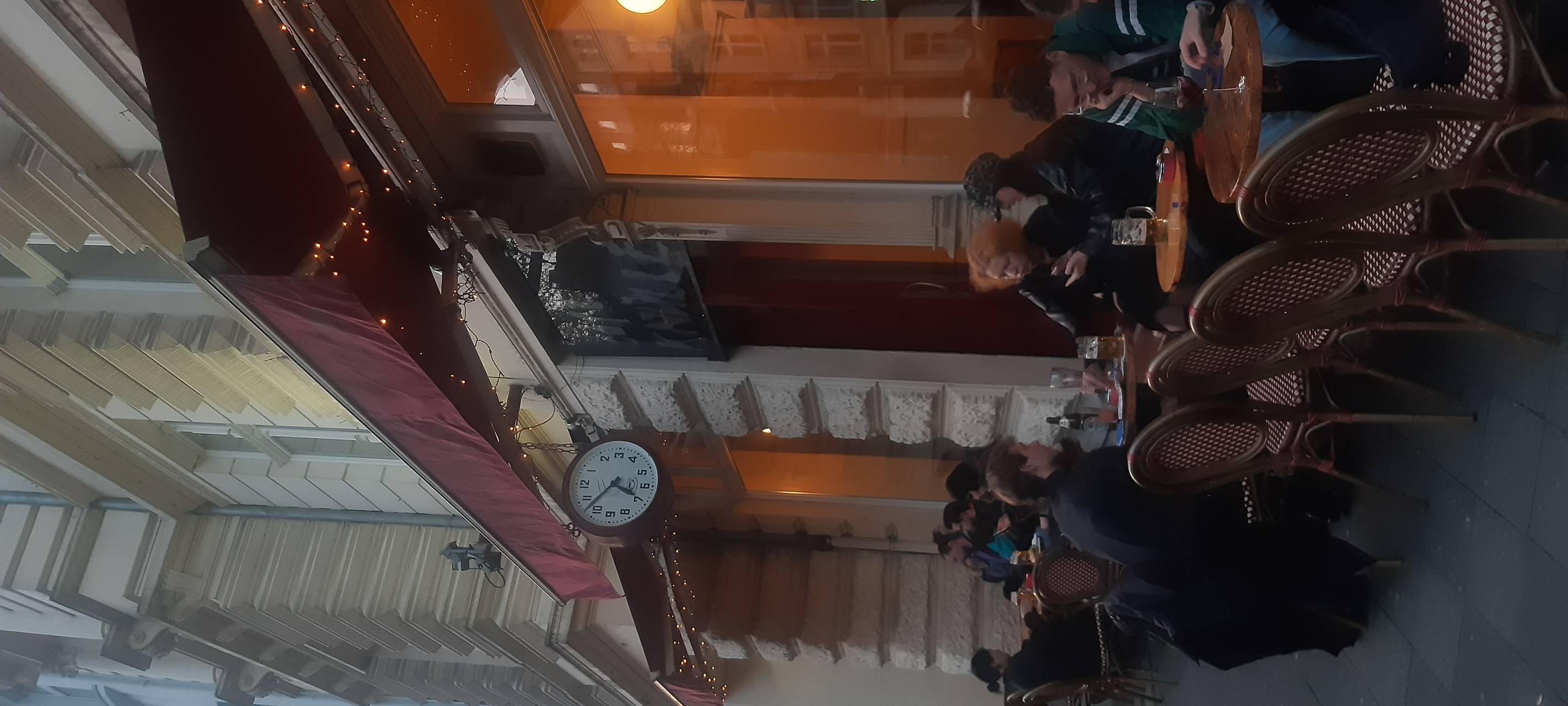 Menschen an kleinen Tischen im Dämmerlicht außen vor einem Café, die Markise ist ausgefahren, links der Bürgersteig.