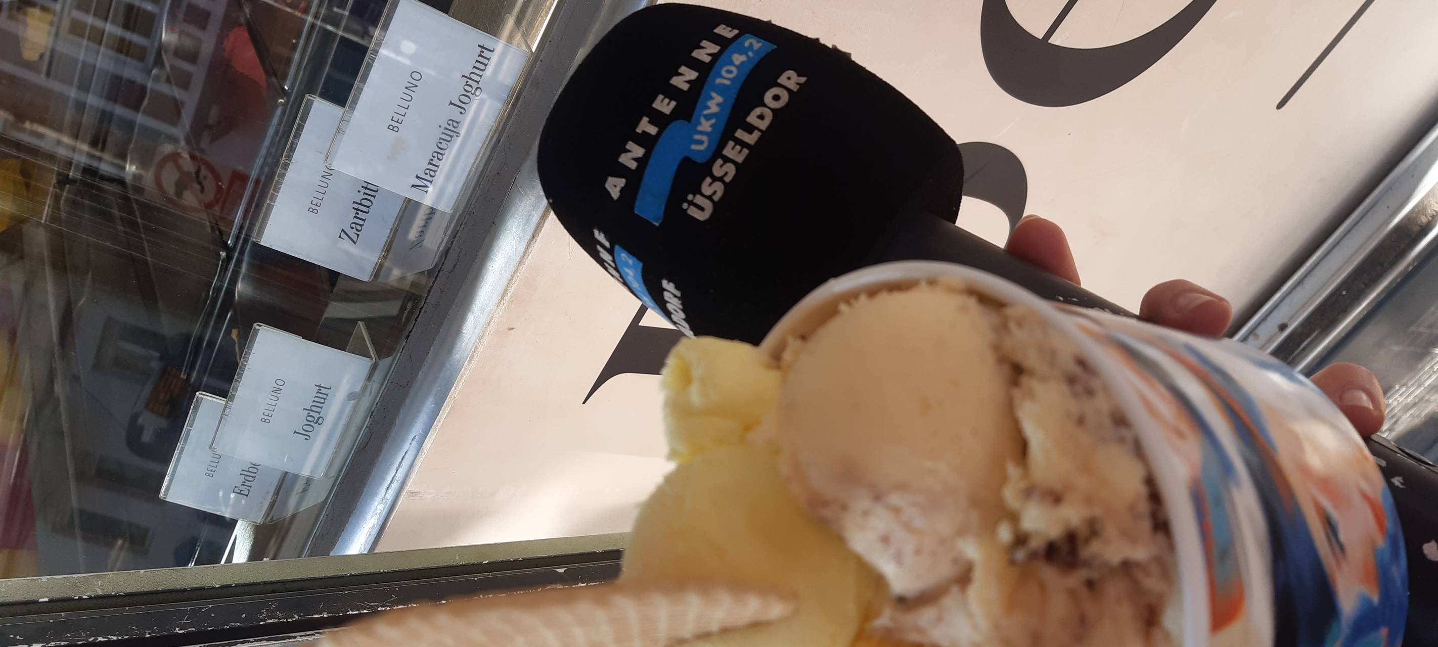 Ein Becher mit mehreren Eiskugeln, daneben das Antenne Düsseldorf Mirkofon, im Hintergrund eine Eistheke