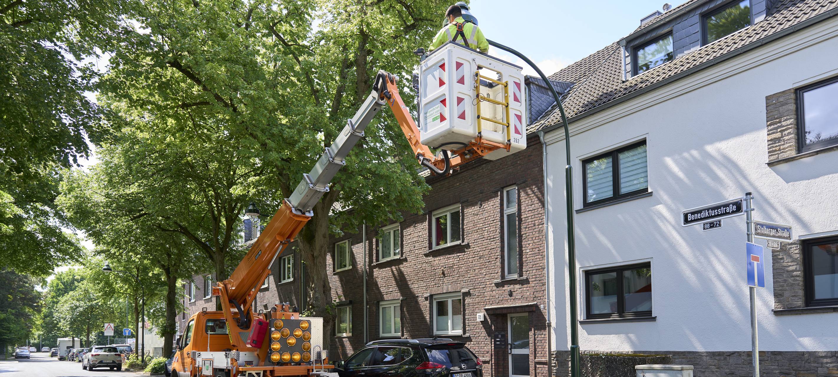 Auf der Benediktusstraße in Heerdt steht ein Kranwagen, von dem aus ein Mitarbeiter der Netzgesellschaft eine Gaslaterne umrüstet. Im Hintergrund sind Wohnhäuser und Bäume zu sehen.