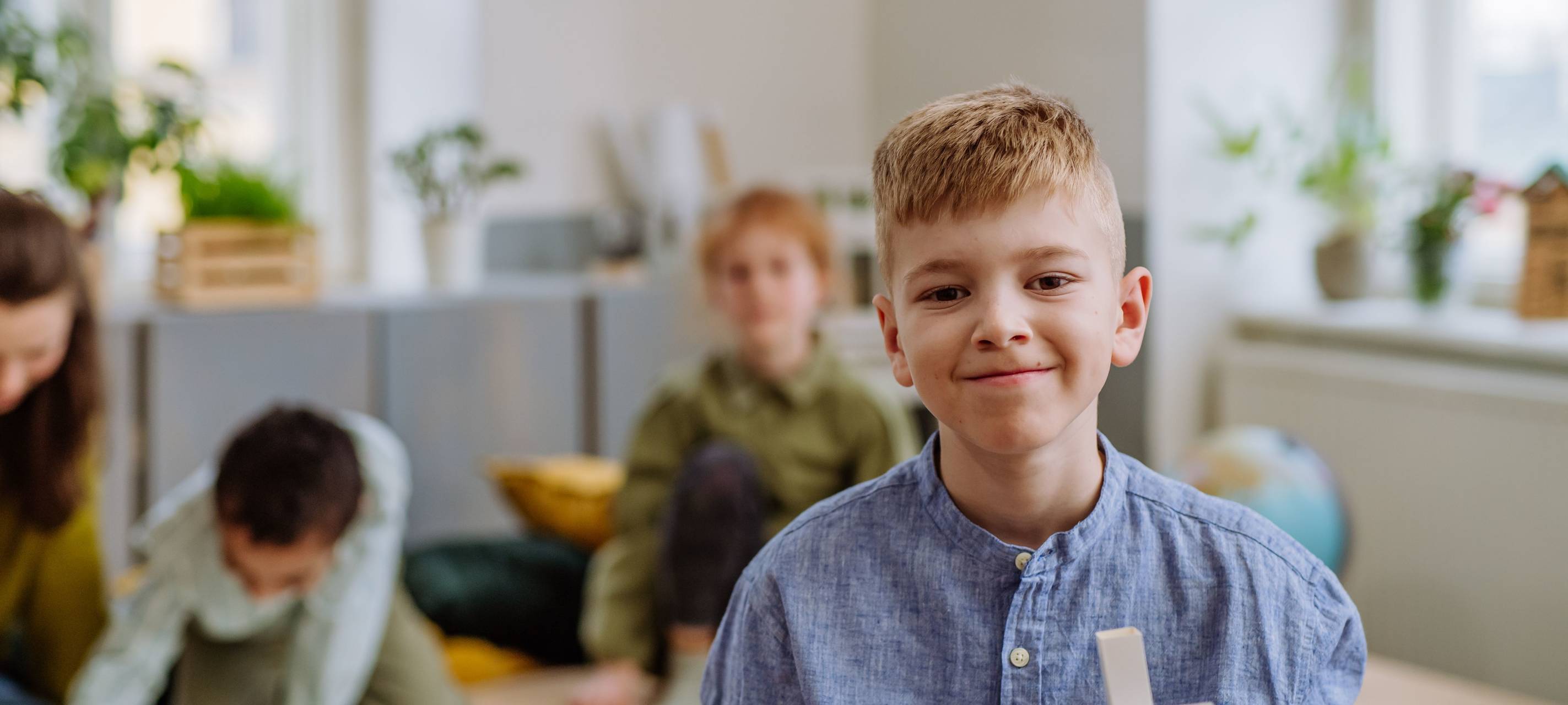 Ein Junge hält ein gebasteltes Holzhaus mit Solaranlagen auf dem Dach in die Kamera und lächelt. Im Hintergrund weitere Schulkinder, die an einem Tisch an etwas arbeiten.