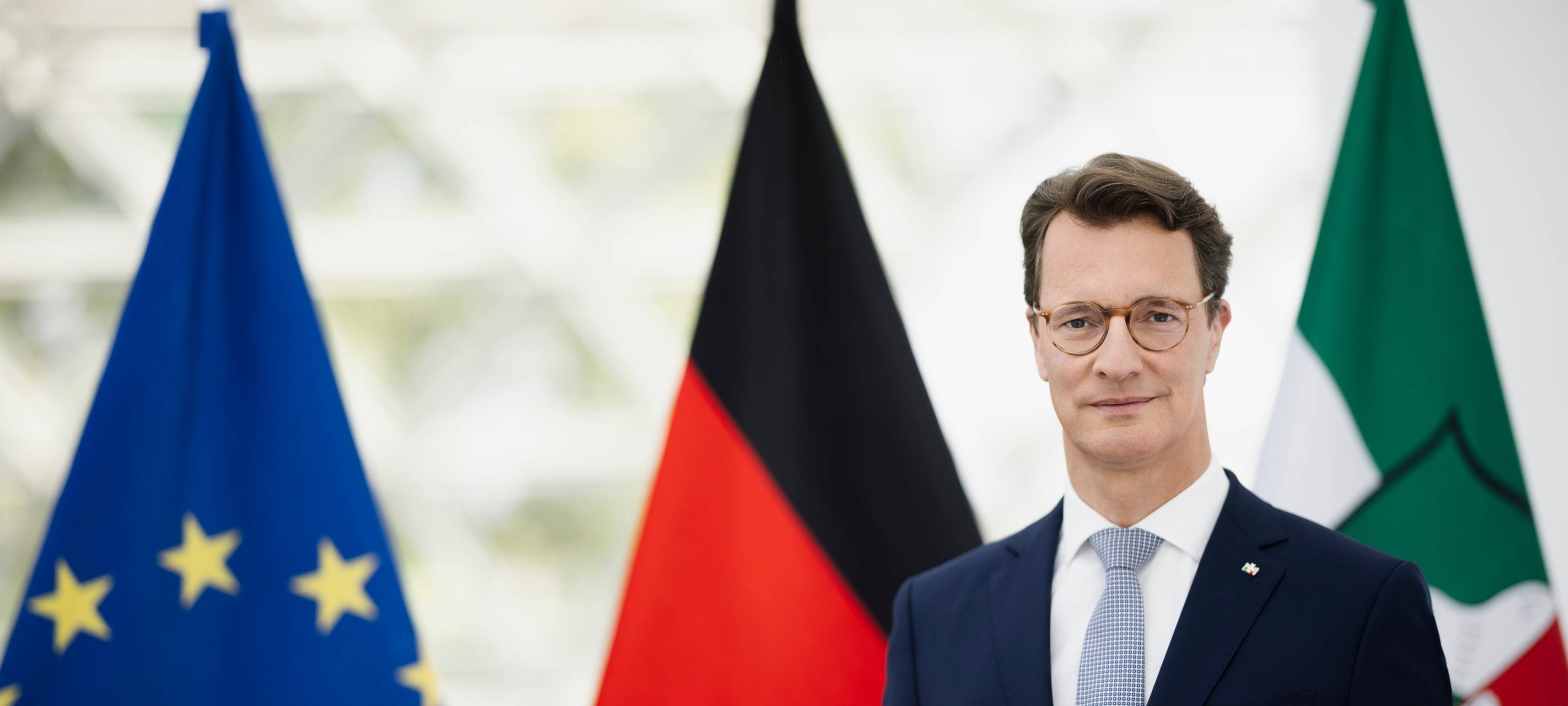 NRW-Ministerpräsident Hendrik Wüst steht vor drei Fahnen (Europa, NRW & Deutschland).