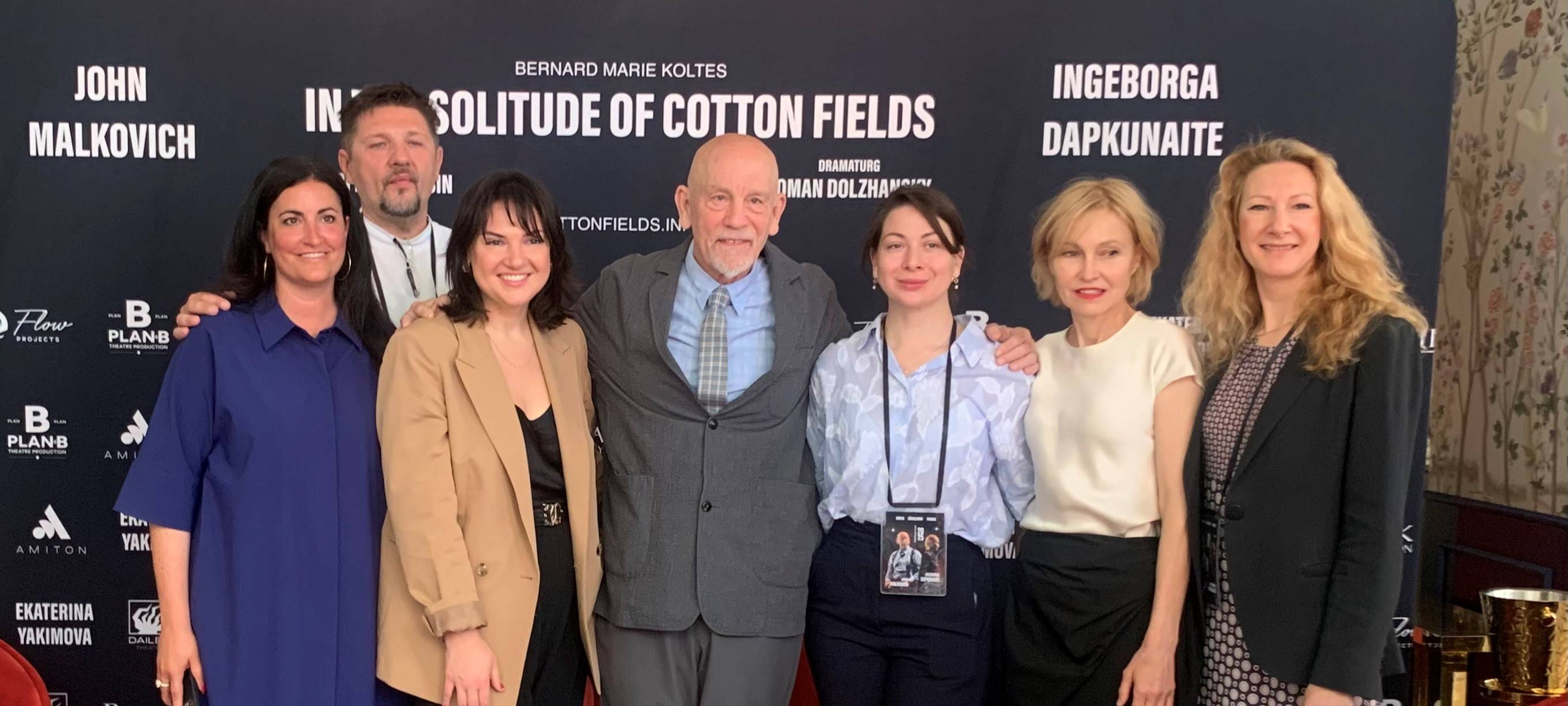 7 Menschen posieren Arm in Arm für ein Foto, in der Mitte John Malkovich. Sie stehen vor einer Werbewand für das Stück In the soltitude of Cotton Fields.