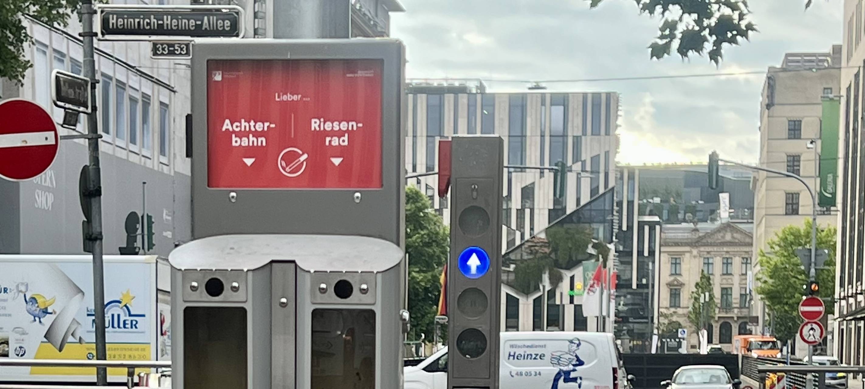 Vor dem Abgang zum U-Bahnhof "Heinrich-Heine-Allee" steht der neue Abstimm-Aschenbecher mit der Frage "Achterbahn oder Riesenrad?". Im Hintergrund ist unter anderem der Kö-Bogen zu sehen.