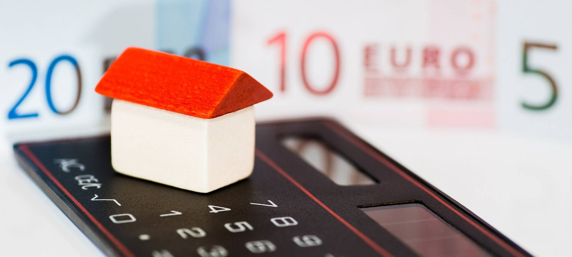 Ein kleines, rot-weißes Haus steht auf einem schwarzen Taschenrechner. Im Hintergrund sind Geldscheine zu sehen.