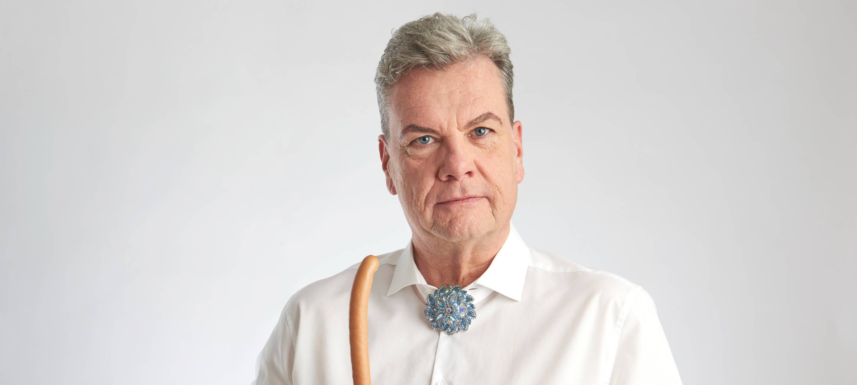 Manes Meckenstock trägt ein weißes Hemd mit einer Brosche und hält ein Bockwürstchen in der rechten Hand.