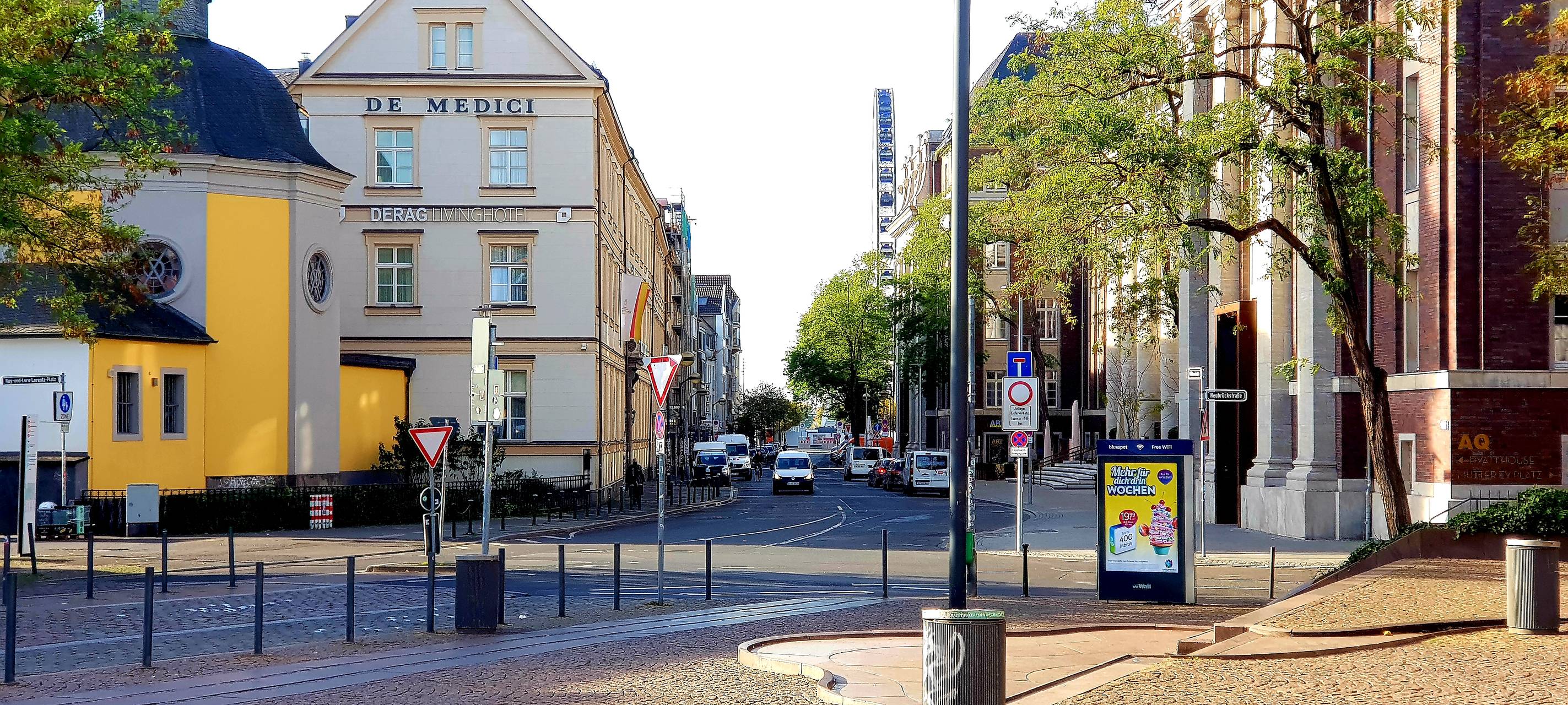Ein städtischer Straßenzug mit einem gelb-weißen Gebäude, geparkten Autos und einem Riesenrad im Hintergrund; rechts ein Backsteingebäude mit Werbetafel.
