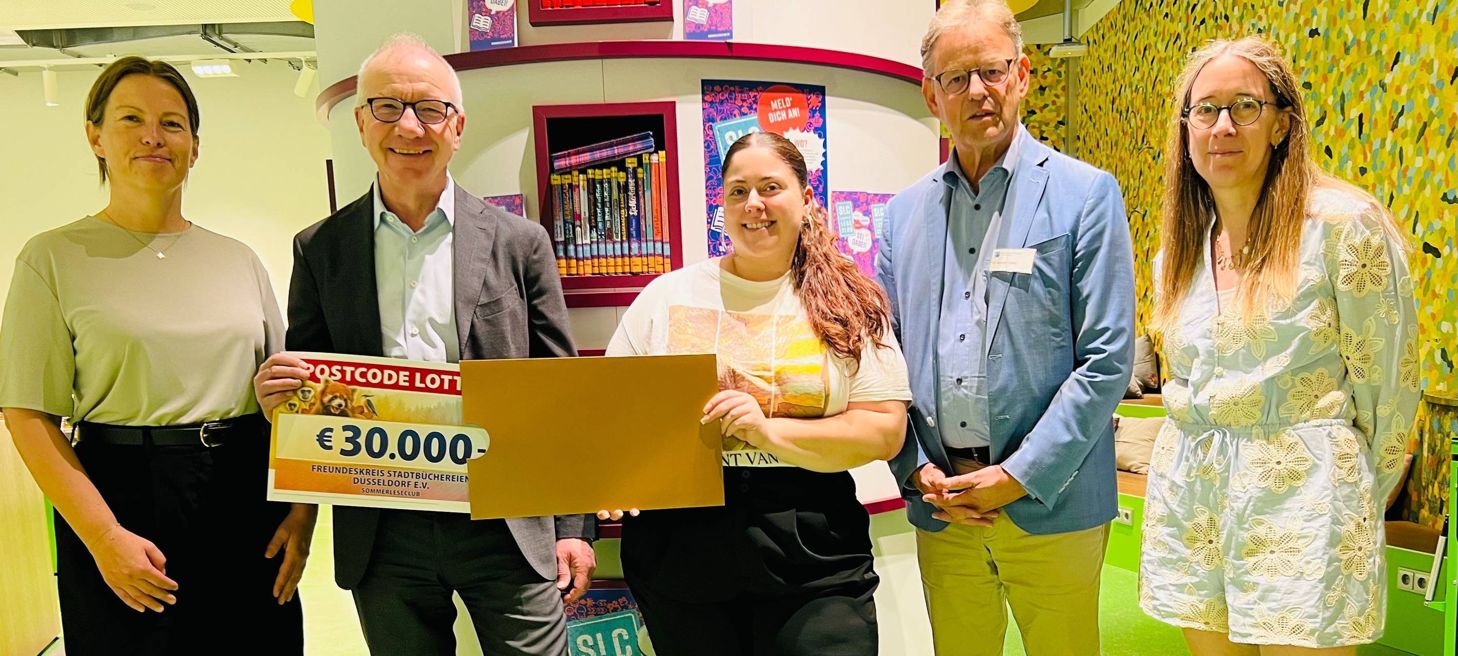 Fünf Menschen stehen in der Stadtbücherei und freuen sich über einen Gutschein über 30.000 Euro der Deutschen Postcode Lotterie