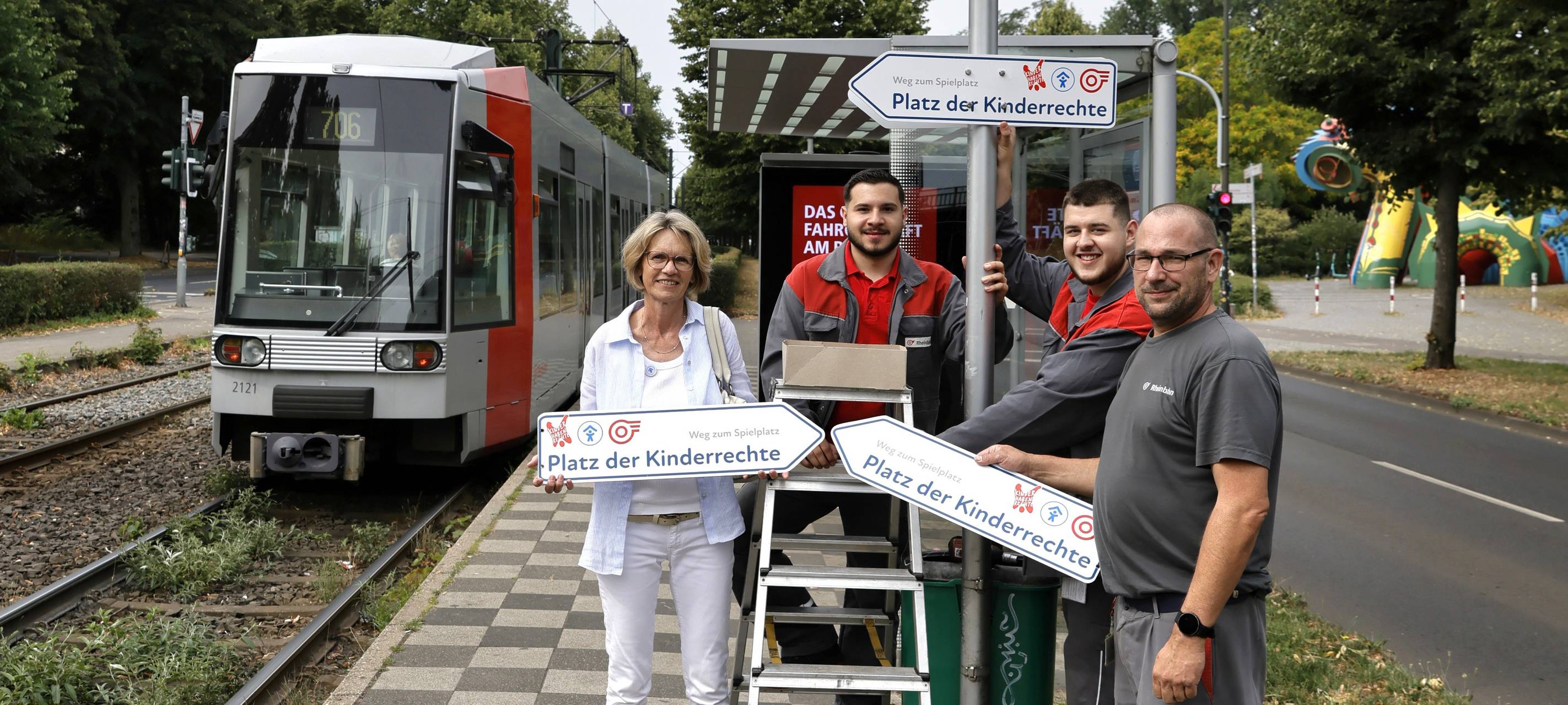 Mitarbeitende von Rheinbahn und Kinderschutzbund mit Schildern (den Platz der) Kinderrechte. Sie stehen neben einer Bahn an der Haltestelle "Volksgarten S".