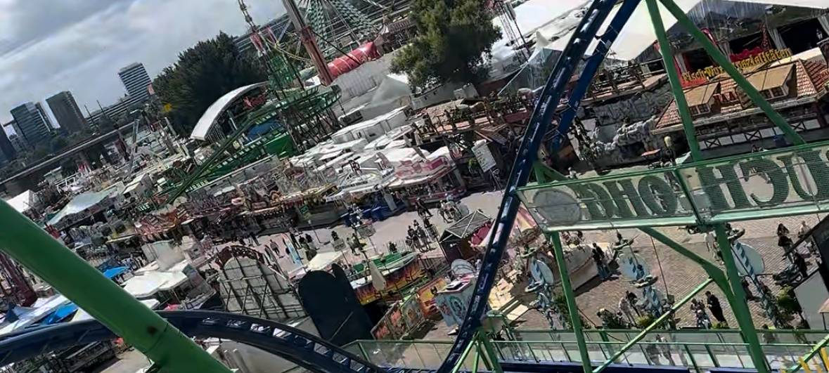 Blick von der Alpina-Achterbahn auf die Rheinkirmes. Im Vordergrund ein Wagen der Alpina-Bahn, im Hintergrund der Kirmesplatz mit Riesenrad und Rheinturm.