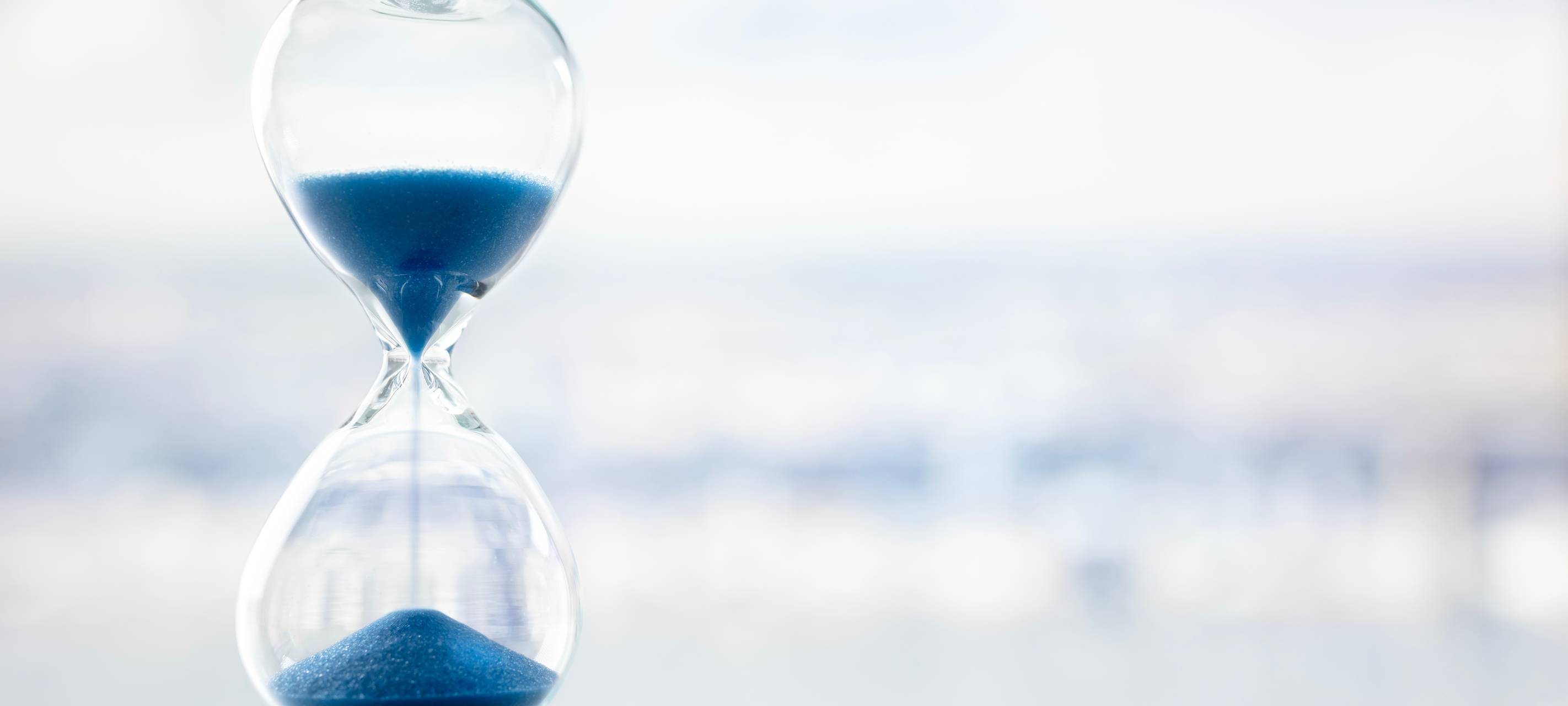 Eine Sanduhr mit blauem Sand vor einem grau-weißen milchigen Hintergrund.