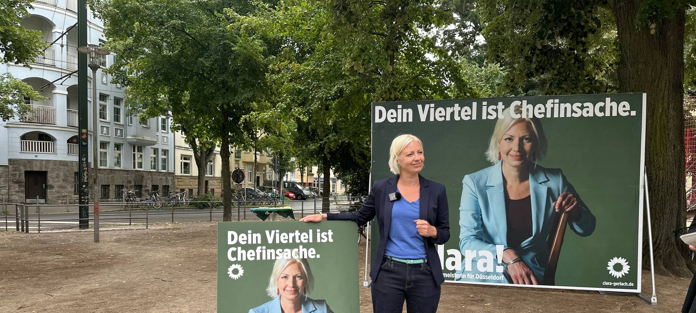 Oberbürgermeister-Kandidatin Clara Gerlach steht zwischen ihren Wahlplakaten, auf denen jeweils Clara Gerlach zu sehen ist. Links ein Laternenplakat, rechts ein Großplakat.