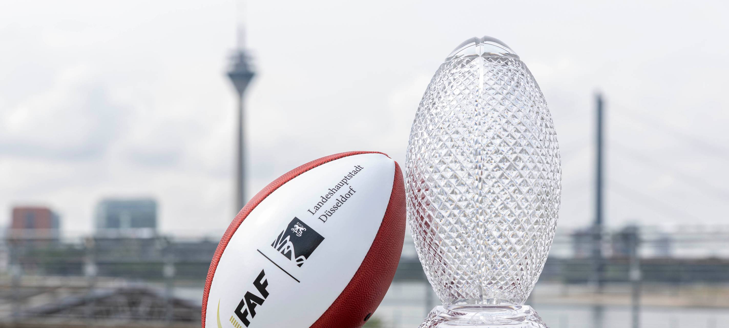 Ein Football und der Pokal der Flag Football WM stehen auf dem Burgplatz. Im Hintergrund sind die Rheinkniebrücke und der Rheinturm.